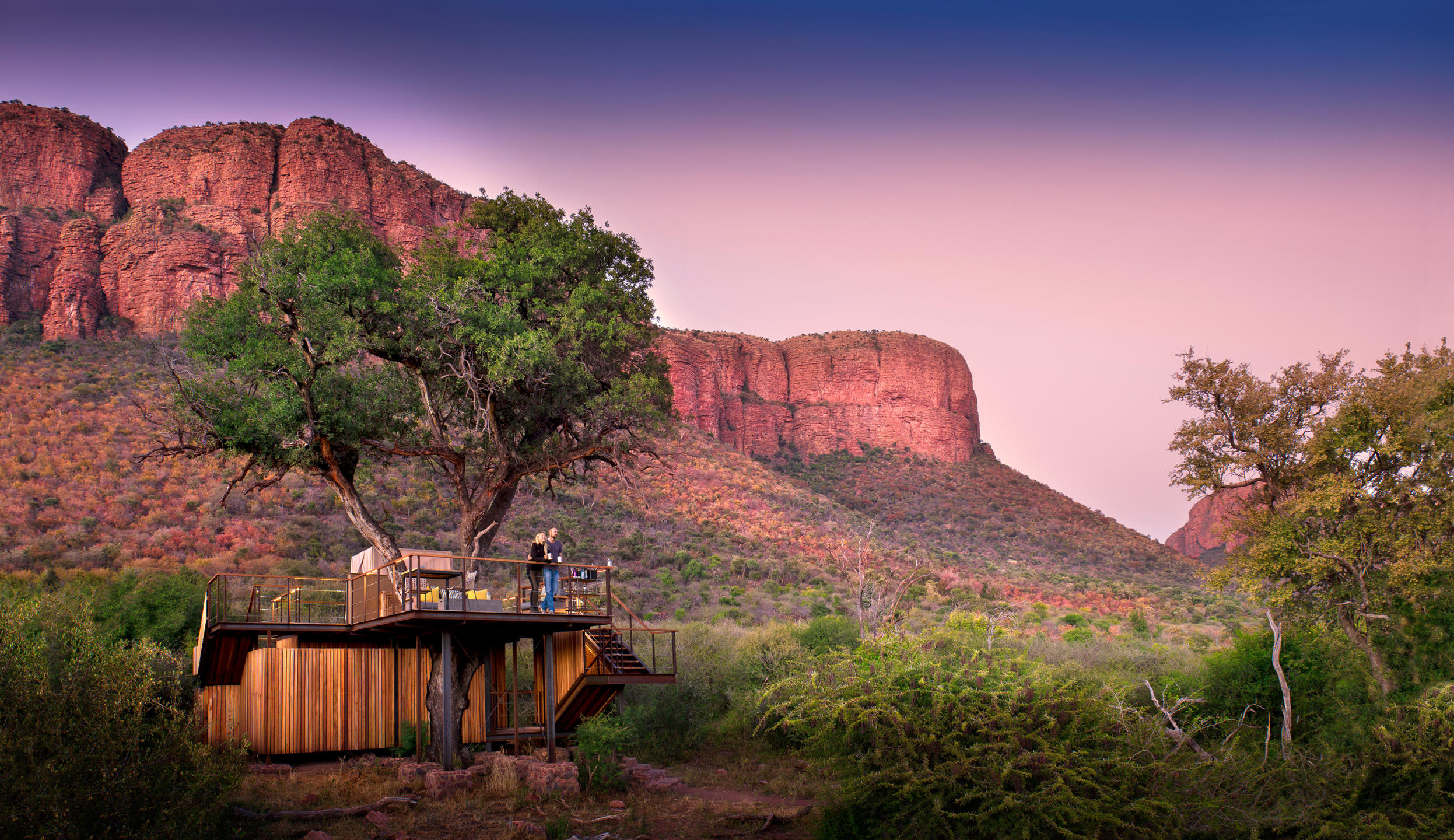 A lit treehouse deck sits beneath towering red cliffs at dusk, with a winding path through dry scrubland.