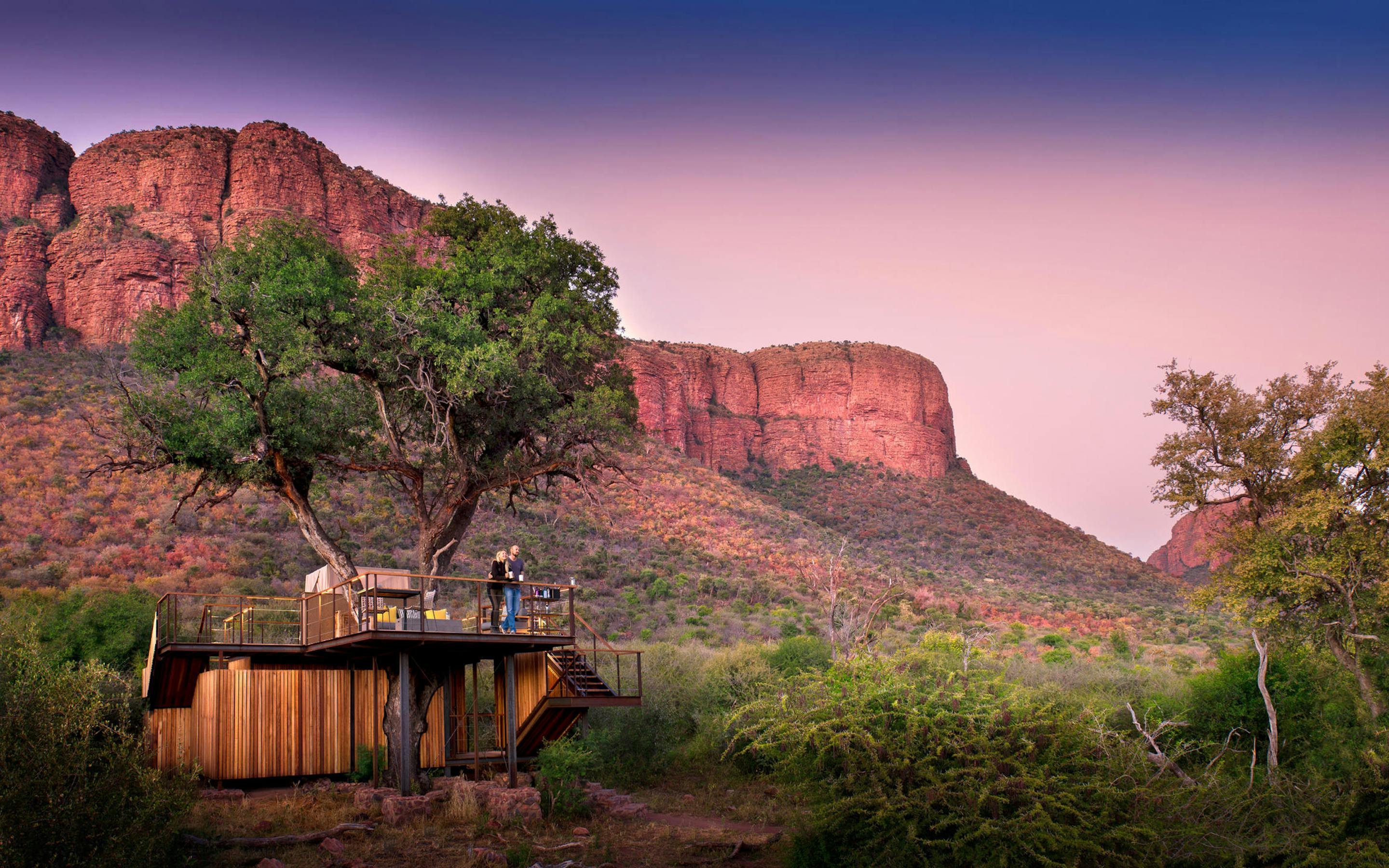 A lit treehouse deck sits beneath towering red cliffs at dusk, with a winding path through dry scrubland.