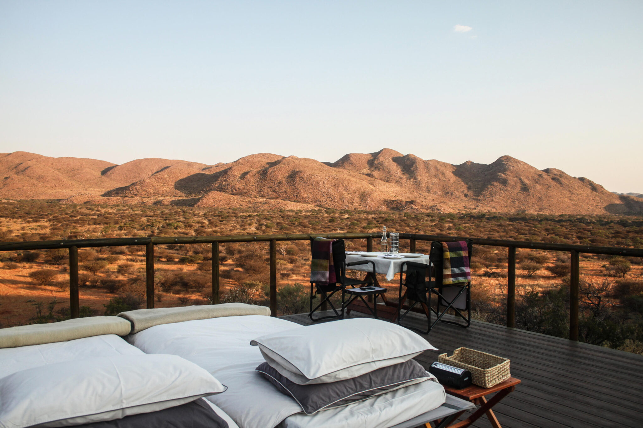 White daybeds and pillows rest on a lodge deck facing red desert hills, with the sun low on the distant horizon.