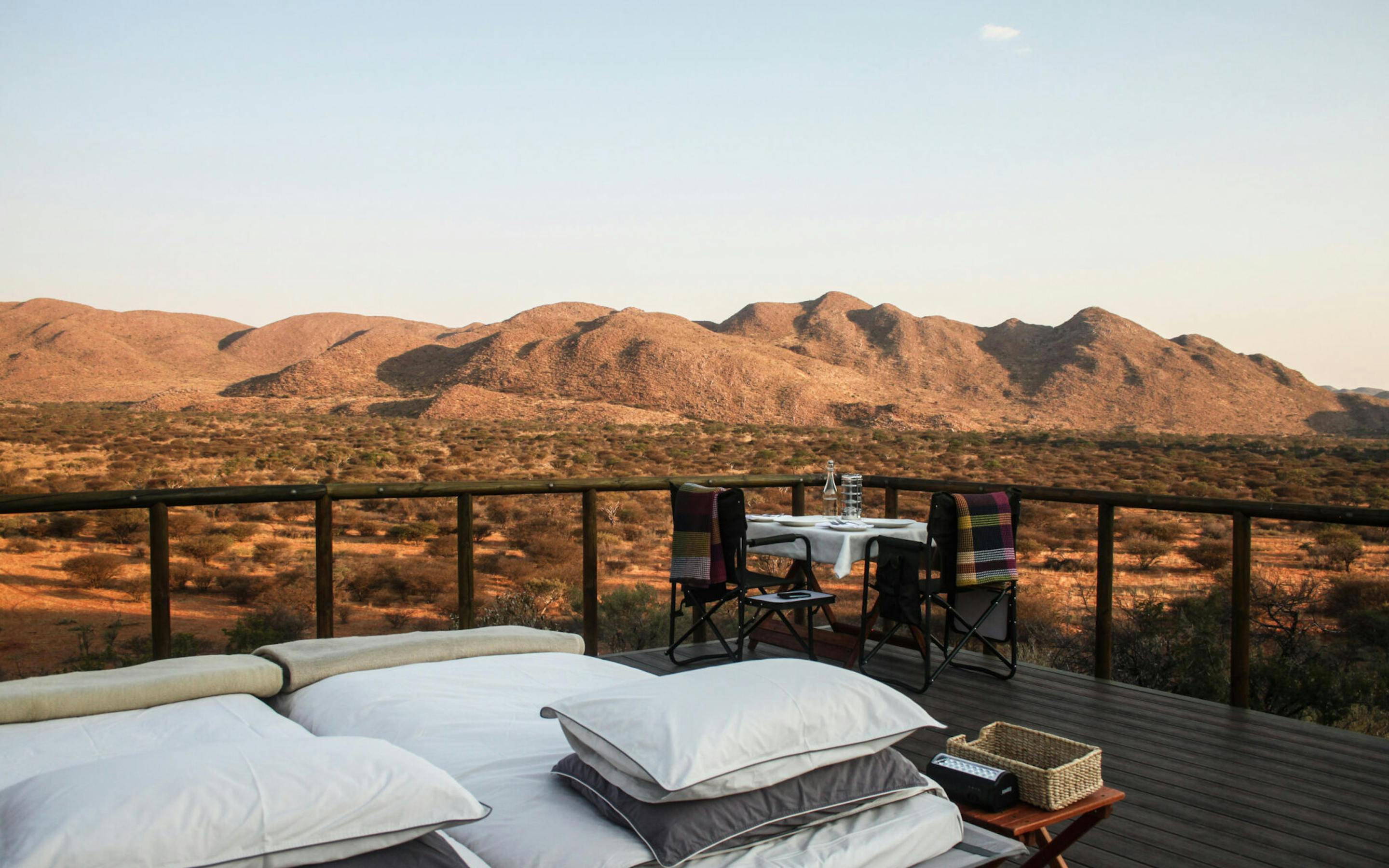 White daybeds and pillows rest on a lodge deck facing red desert hills, with the sun low on the distant horizon.