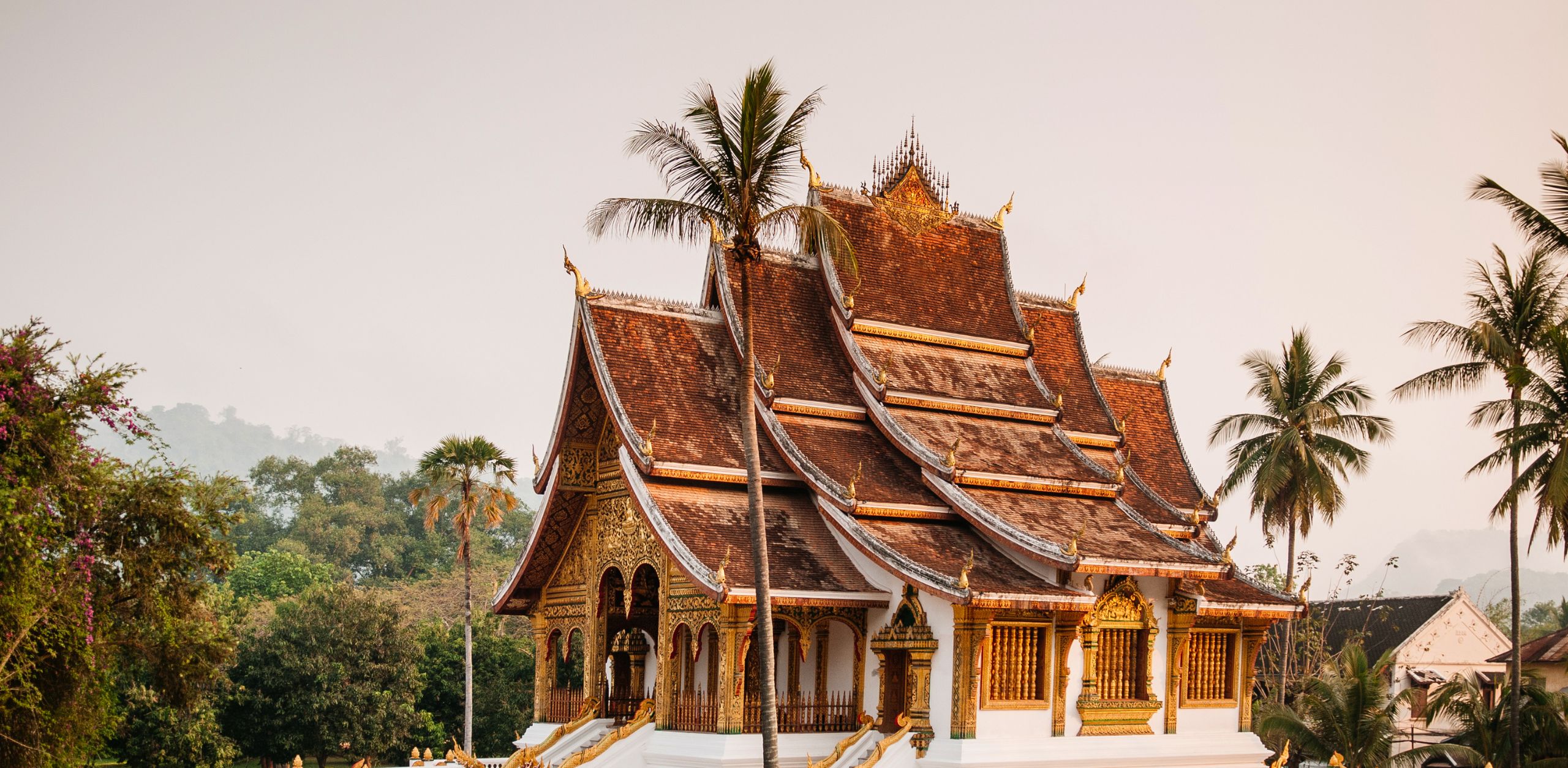 Ornate temple with layered roofs and gilded details rises above palms and lush gardens under a warm hazy sky.