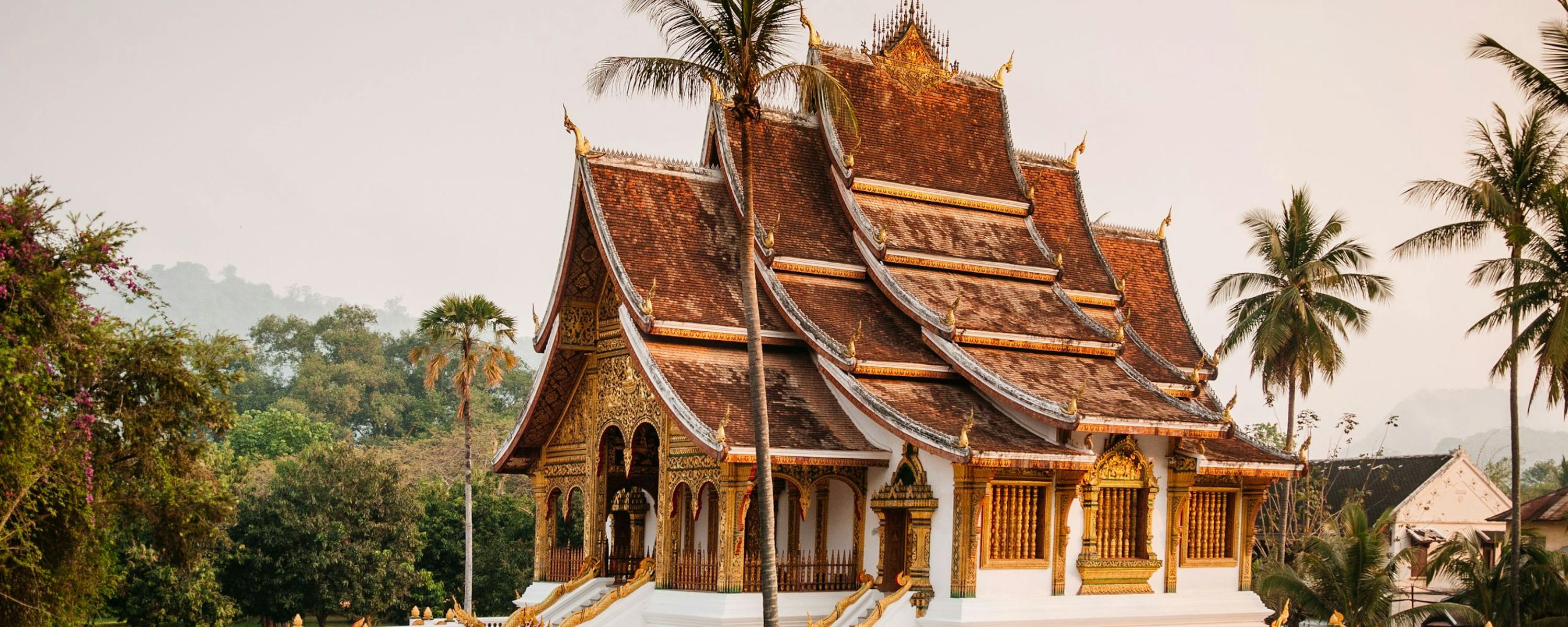 Ornate temple with layered roofs and gilded details rises above palms and lush gardens under a warm hazy sky.