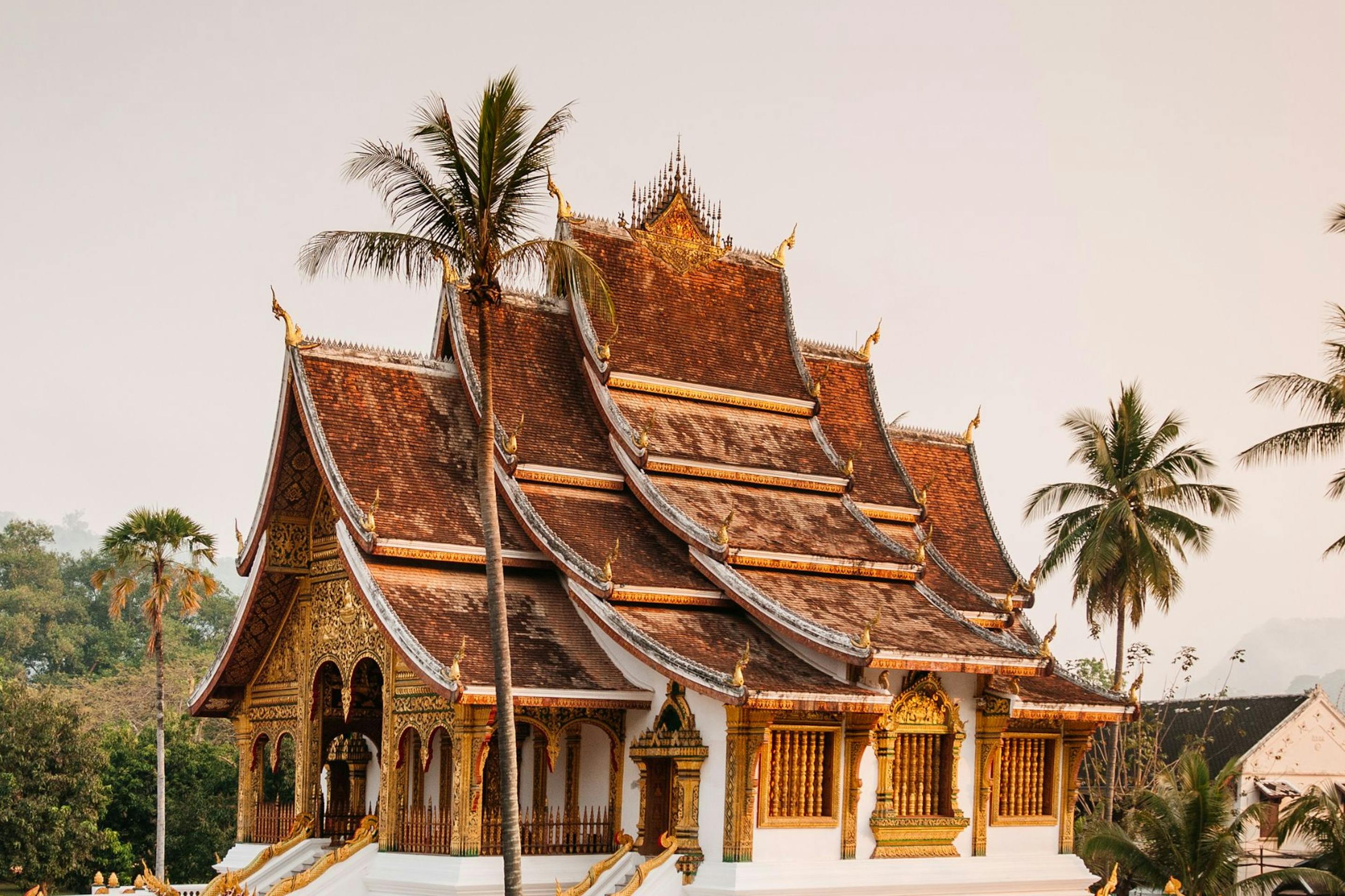 Ornate temple with layered roofs and gilded details rises above palms and lush gardens under a warm hazy sky.