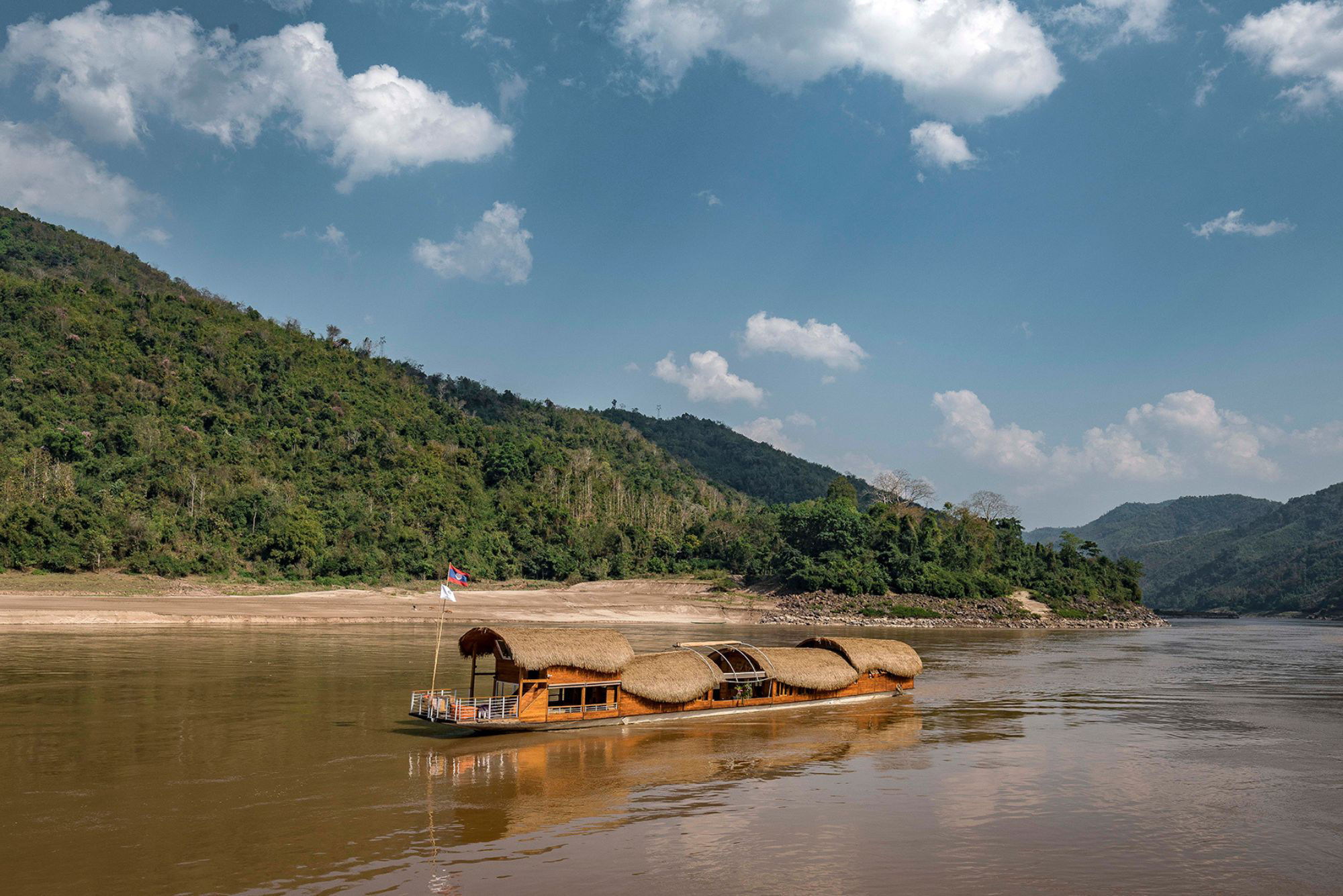 A long wooden boat glides along a wide brown river beside forested hills, reflecting clouds in calm water.