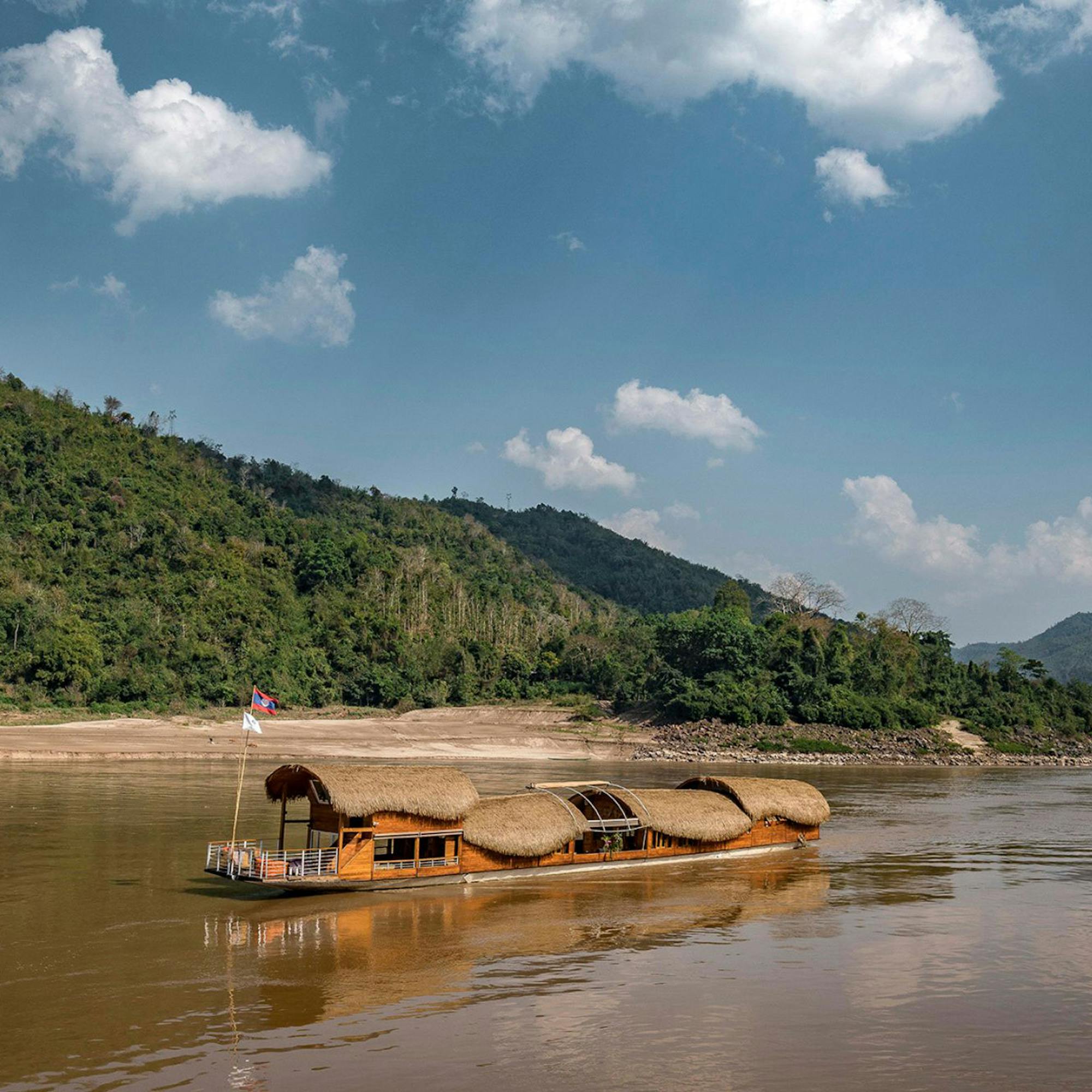 A long wooden boat glides along a wide brown river beside forested hills, reflecting clouds in calm water.