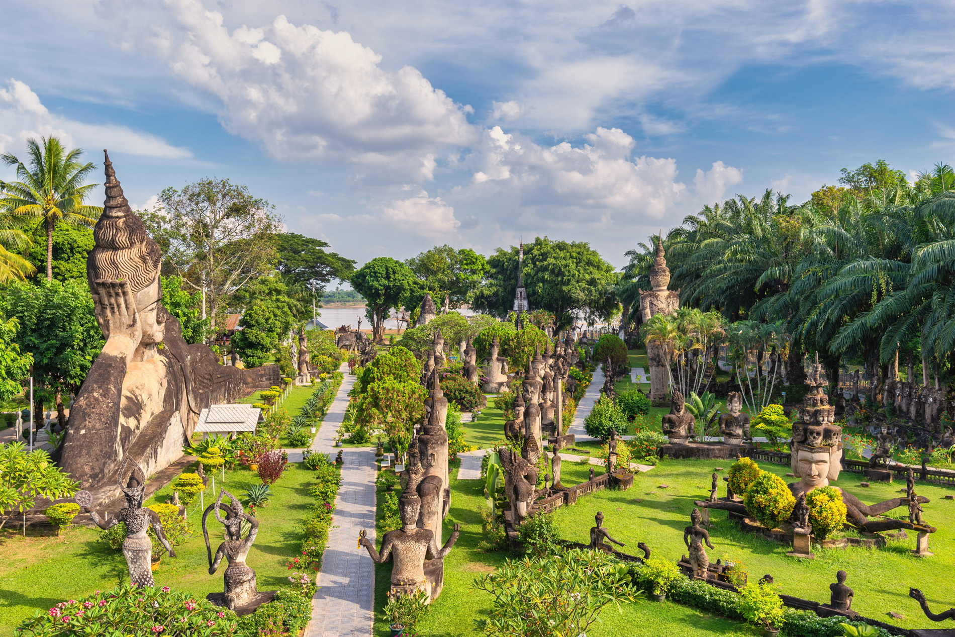 Stone statues line a grassy path in a sculpture garden, with palm trees and bright clouds above the green grounds.