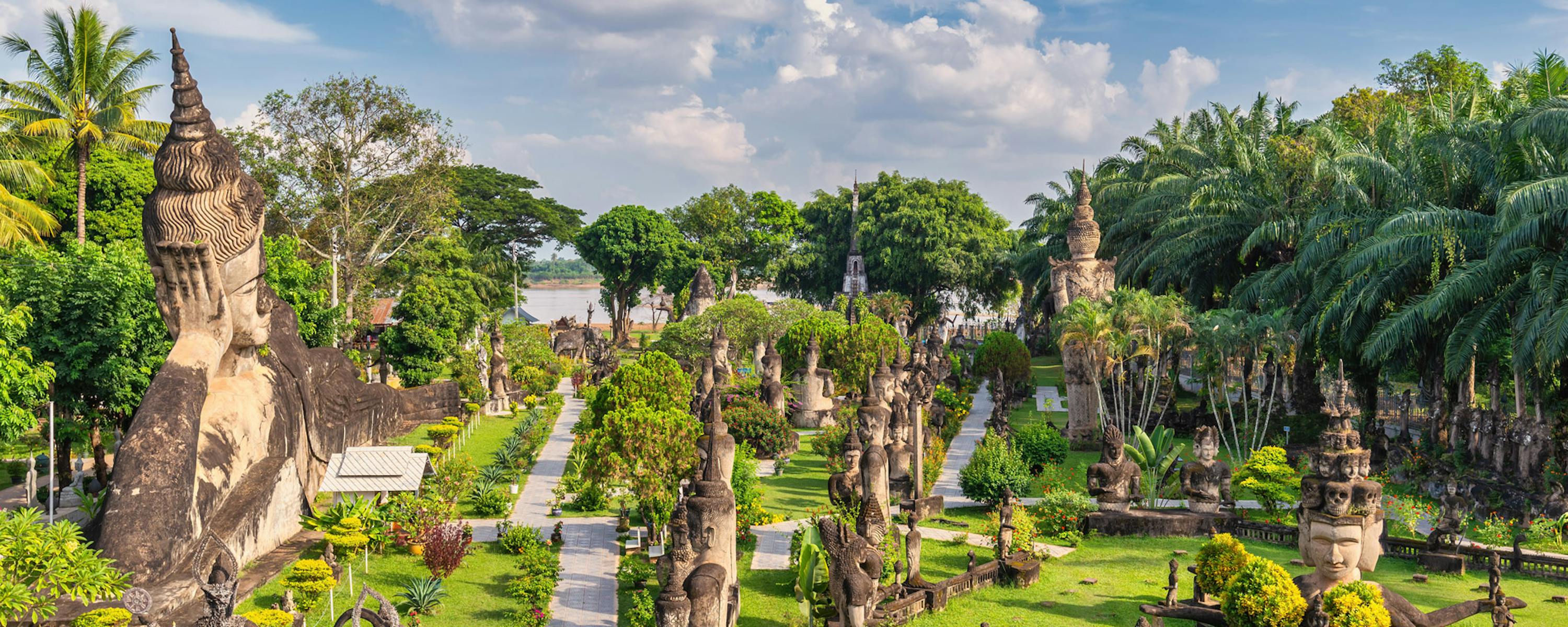 Stone statues line a grassy path in a sculpture garden, with palm trees and bright clouds above the green grounds.