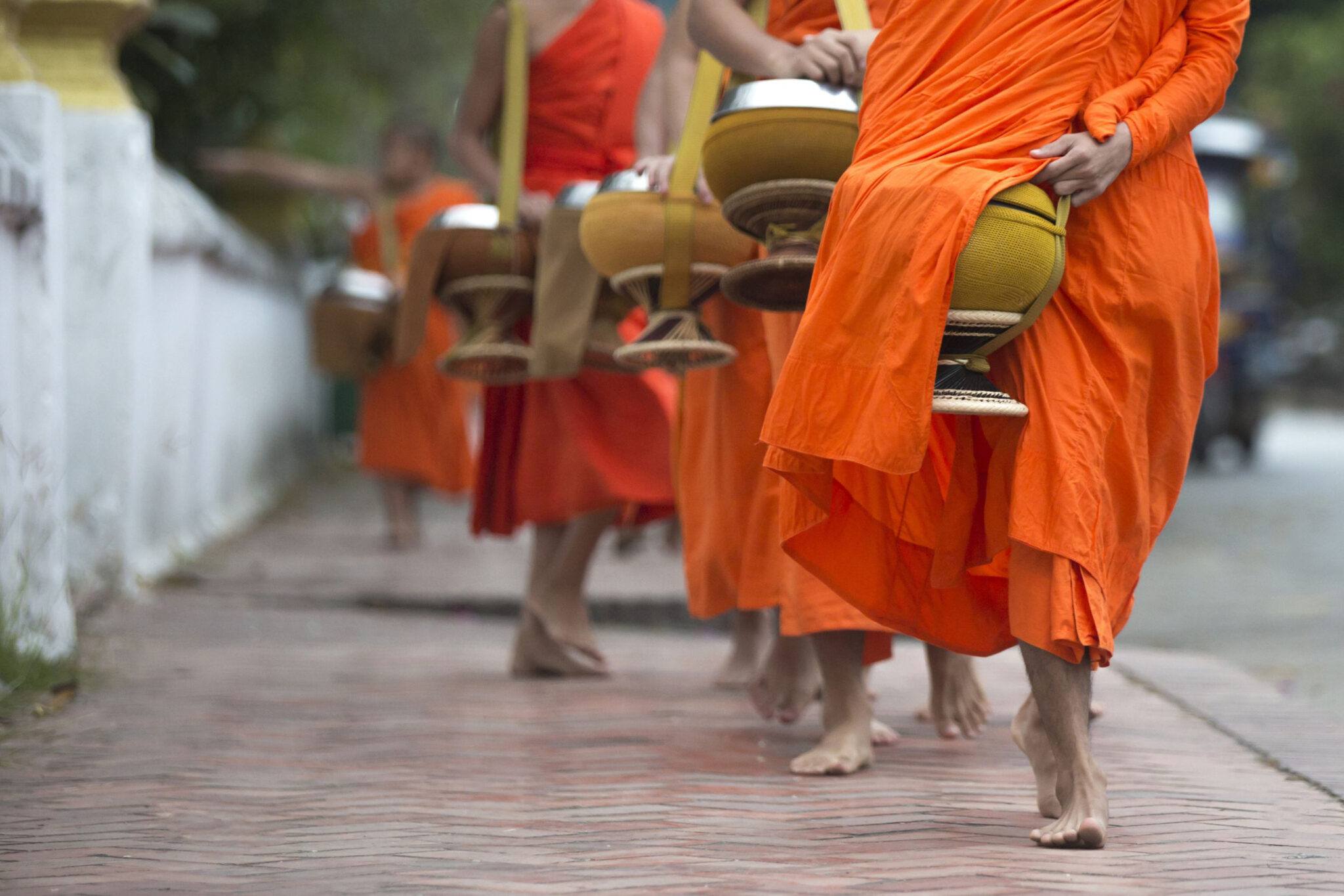 Buddhist monks in orange robes walk along a brick sidewalk, carrying metal bowls during an alms-giving procession.