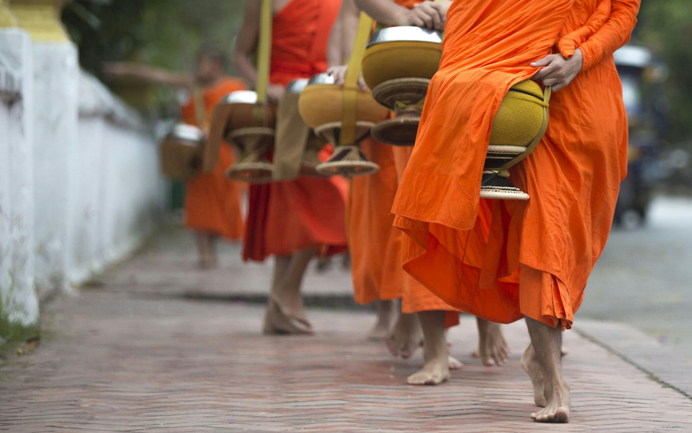 Buddhist monks in orange robes walk along a brick sidewalk, carrying metal bowls during an alms-giving procession.