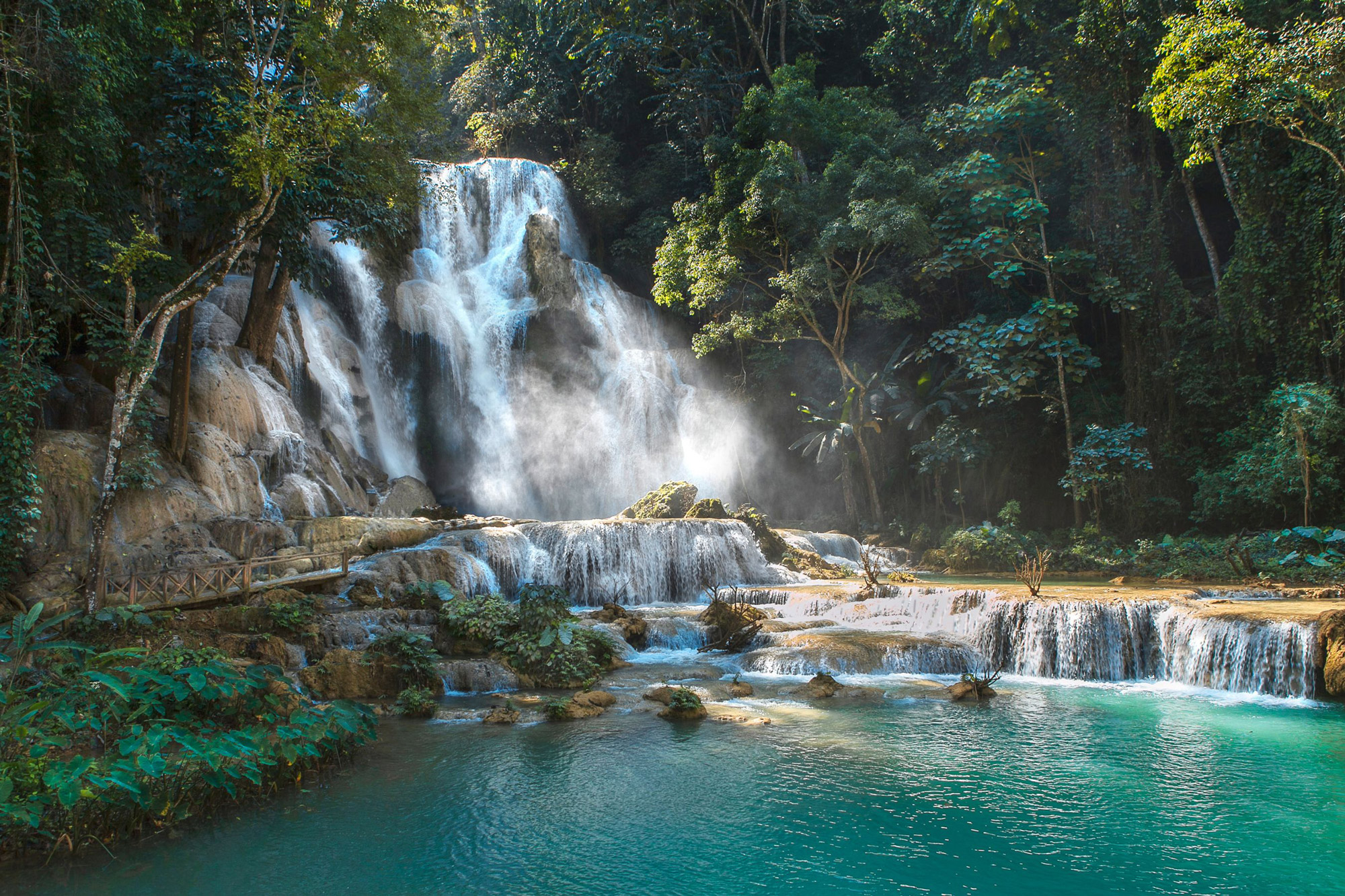 A multi-tiered waterfall spills into bright turquoise pools surrounded by dense jungle foliage and mossy rocks.