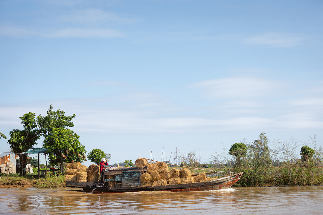 A wooden boat carrying people and cargo moves along a wide river, with grassy banks and pale blue sky above.