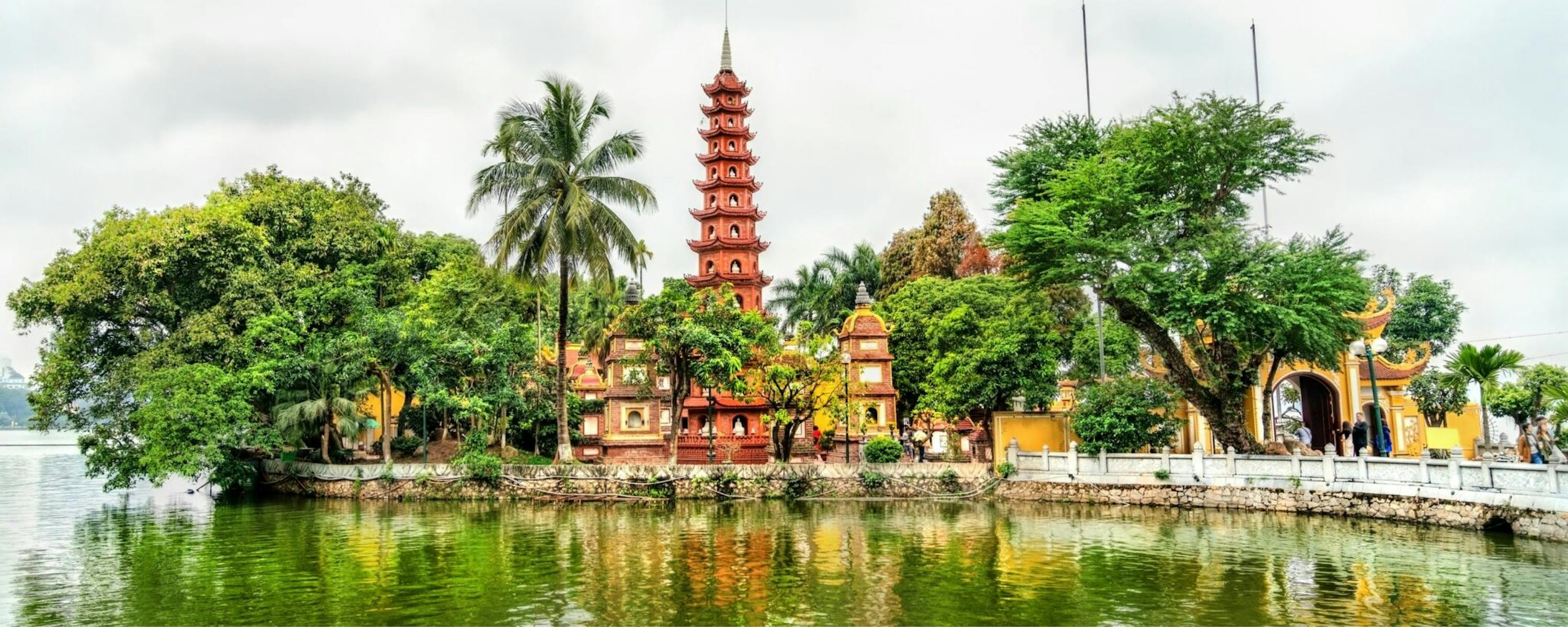 A red pagoda rises beside a calm lake, framed by palms and trees with soft reflections in the water below.