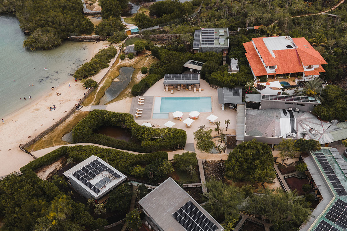 Aerial view of a beachfront hotel with a central pool, sun umbrellas, and paths leading to the sand and bay.
