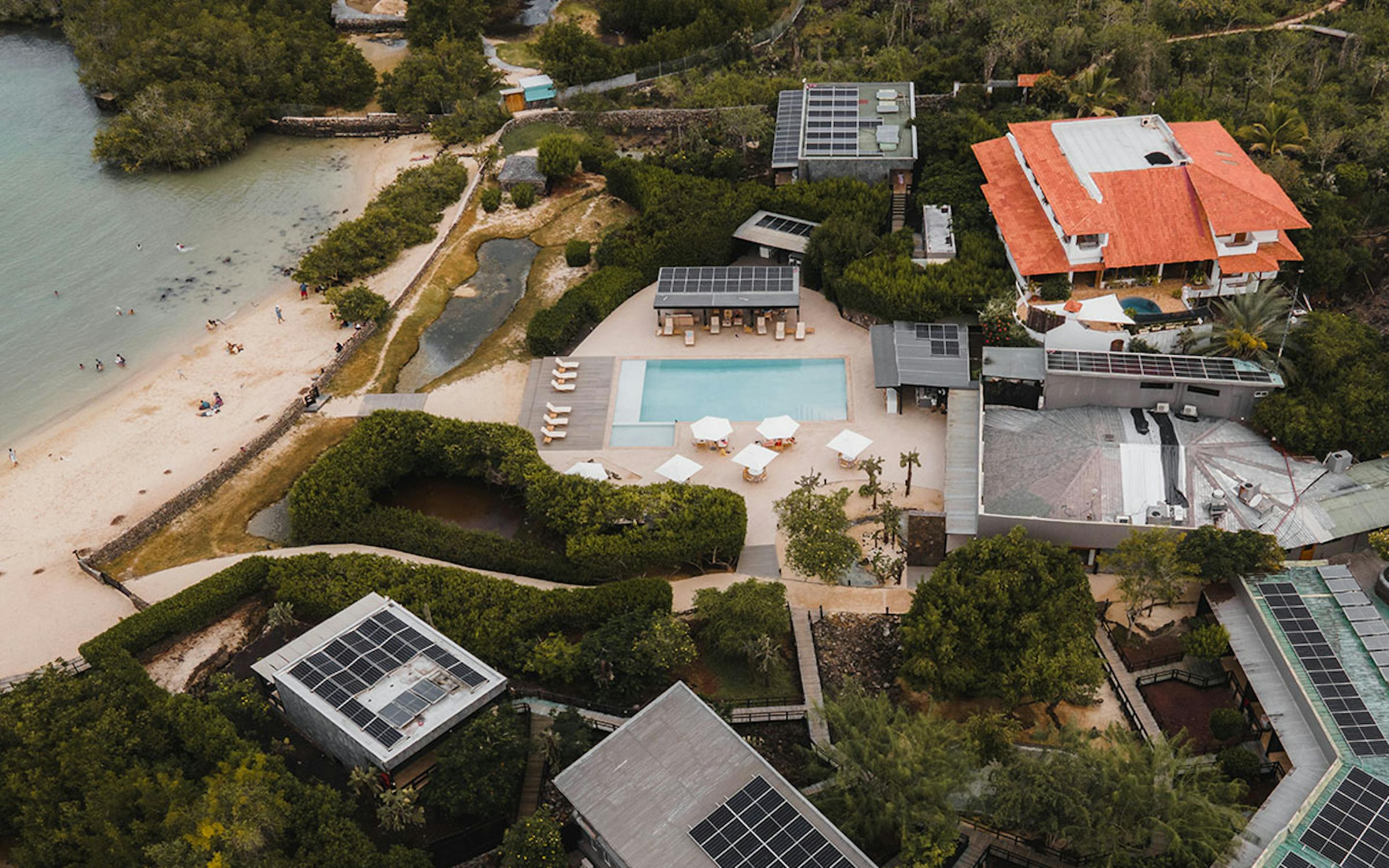 Aerial view of a beachfront hotel with a central pool, sun umbrellas, and paths leading to the sand and bay.