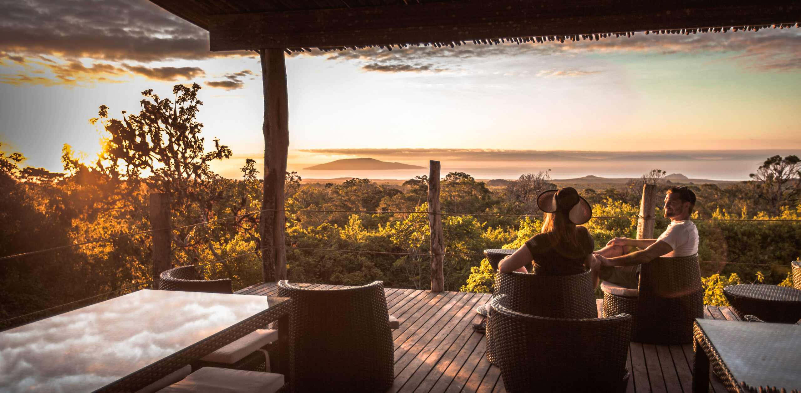 Guests relax on an open deck at sunset, with silhouettes framed by warm light and orange clouds over distant hills.