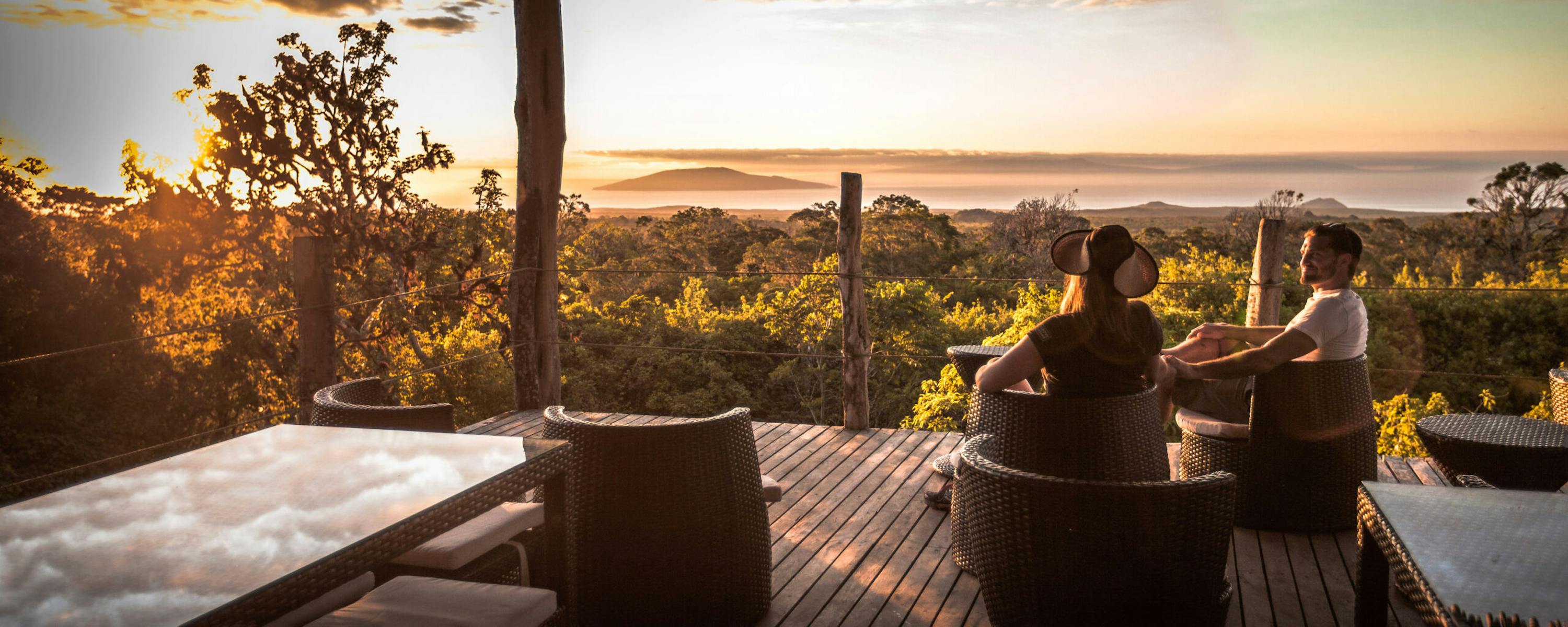 Guests relax on an open deck at sunset, with silhouettes framed by warm light and orange clouds over distant hills.
