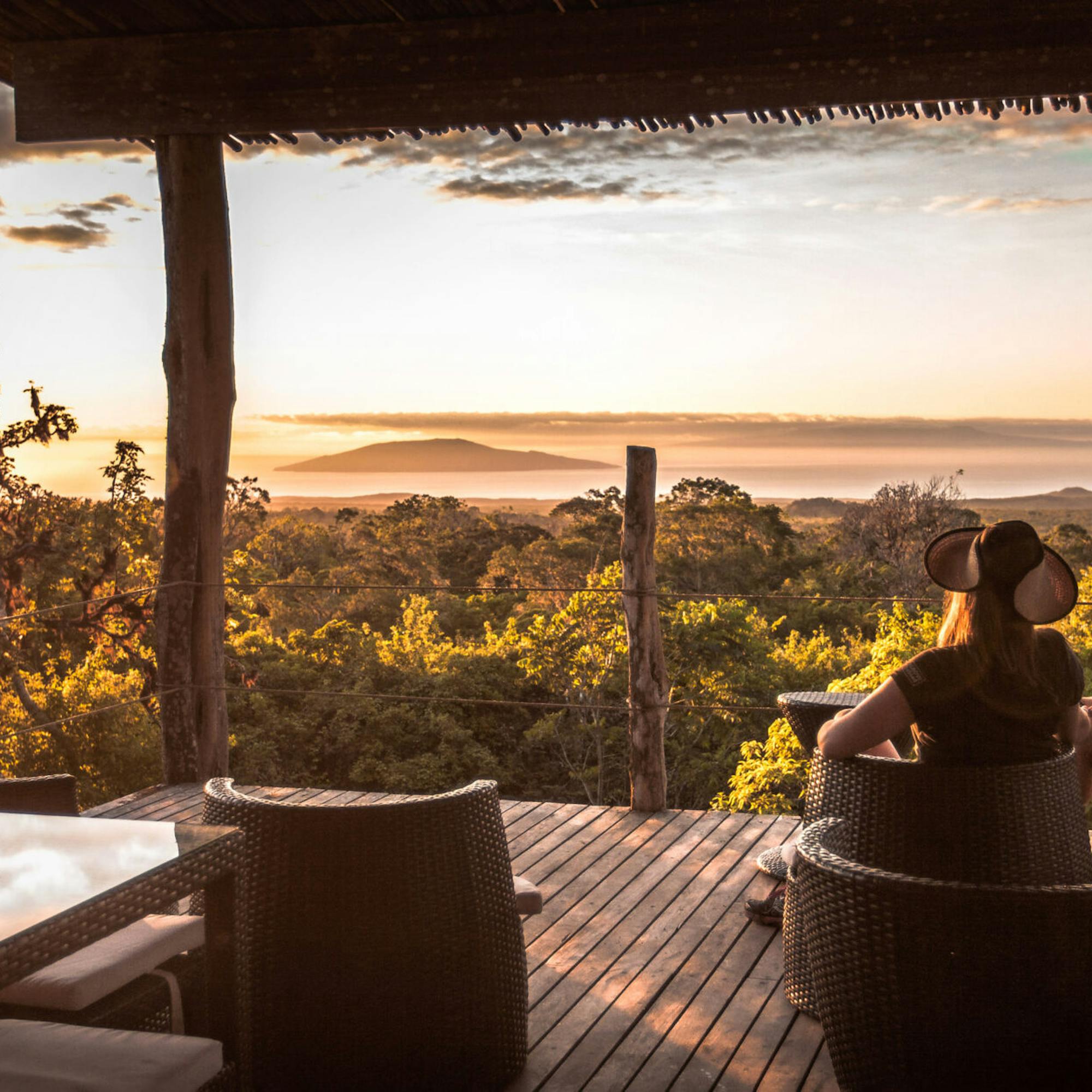 Guests relax on an open deck at sunset, with silhouettes framed by warm light and orange clouds over distant hills.