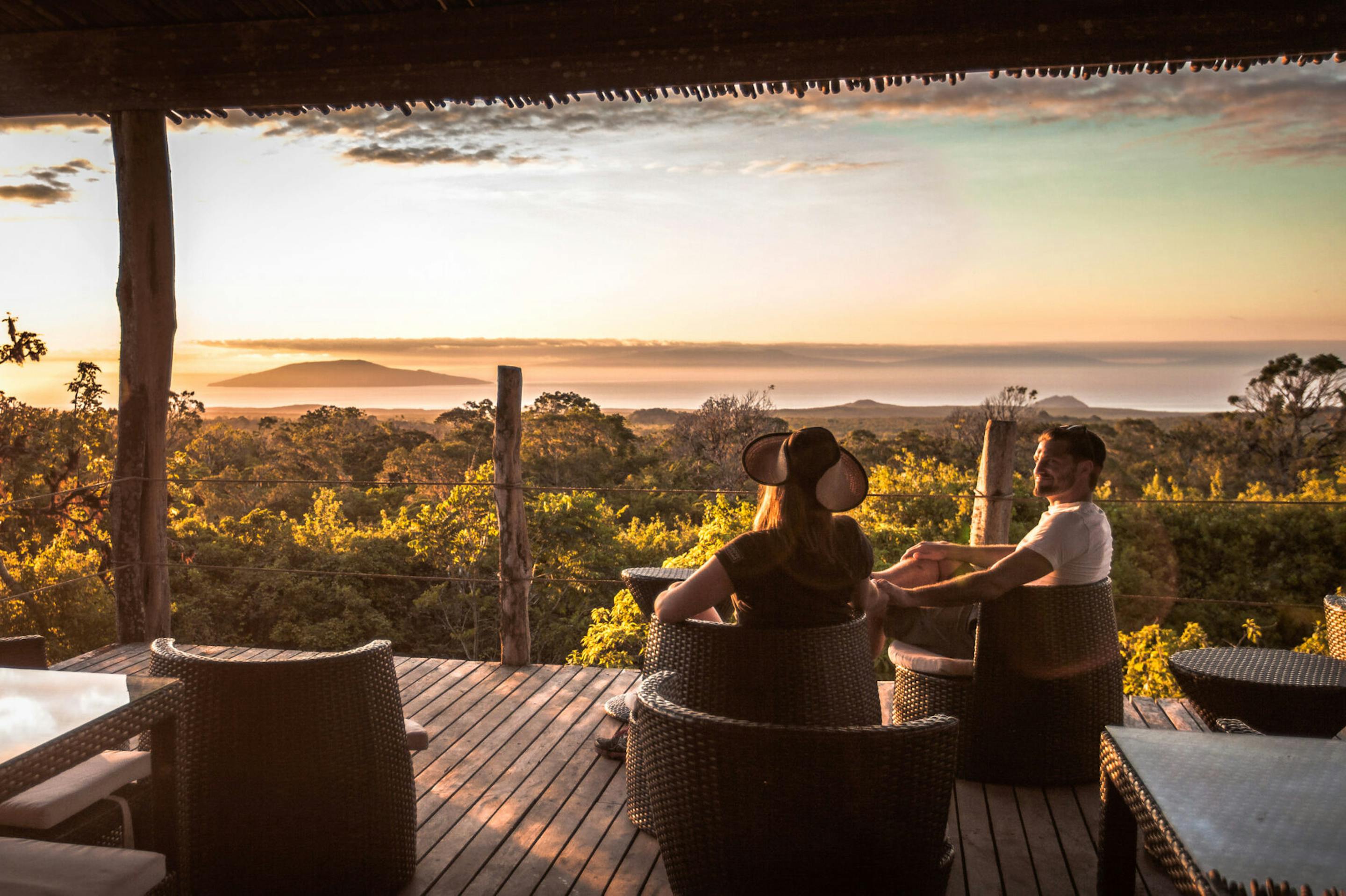 Guests relax on an open deck at sunset, with silhouettes framed by warm light and orange clouds over distant hills.