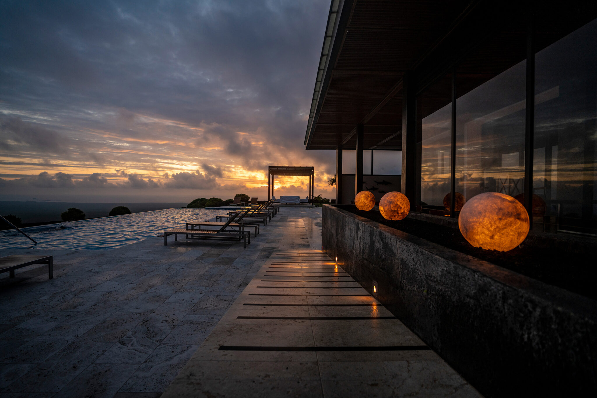 Lantern-lit walkway beside lounge chairs reflects in wet stone as sunset clouds glow beyond a sleek hotel facade.