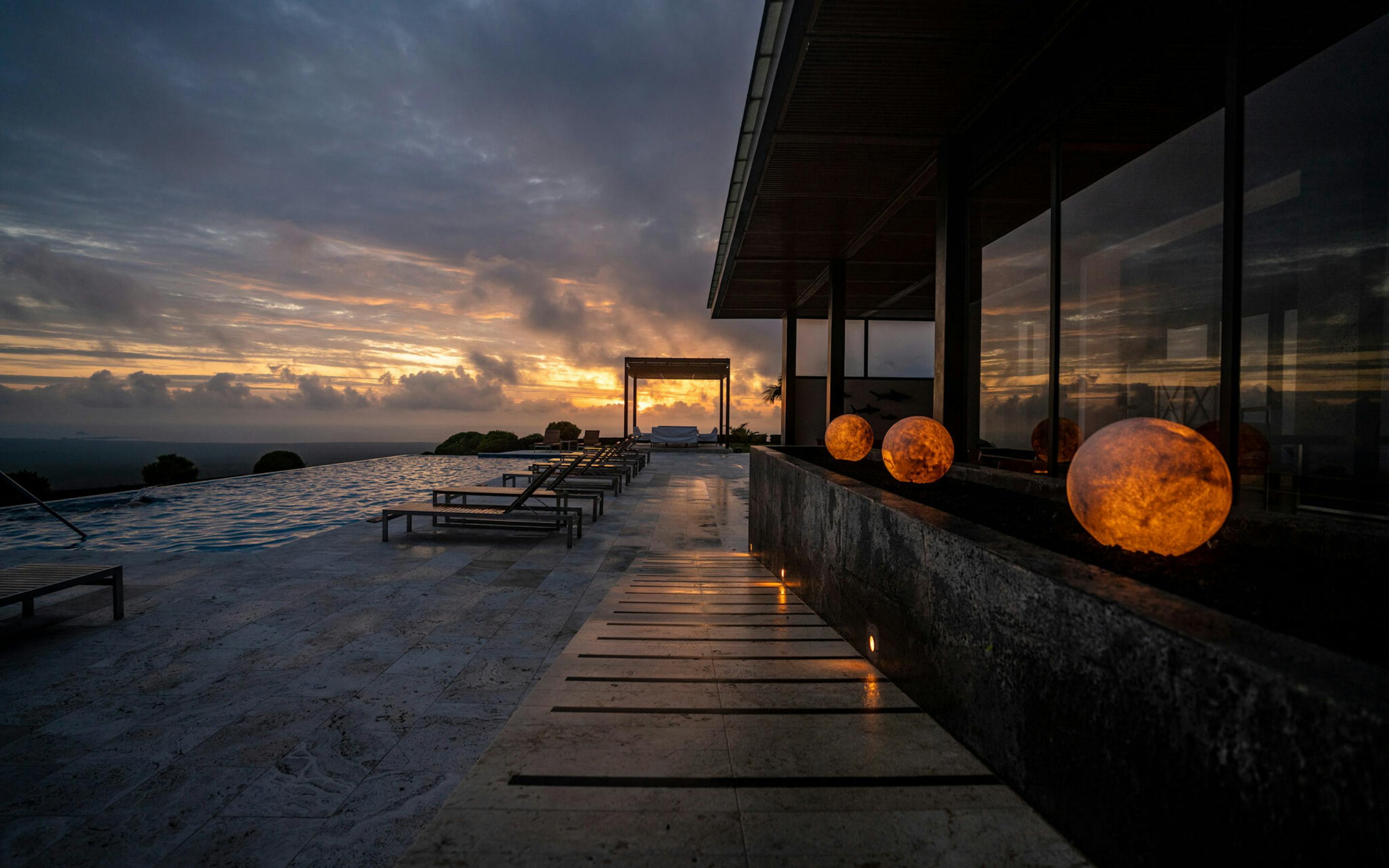 Lantern-lit walkway beside lounge chairs reflects in wet stone as sunset clouds glow beyond a sleek hotel facade.