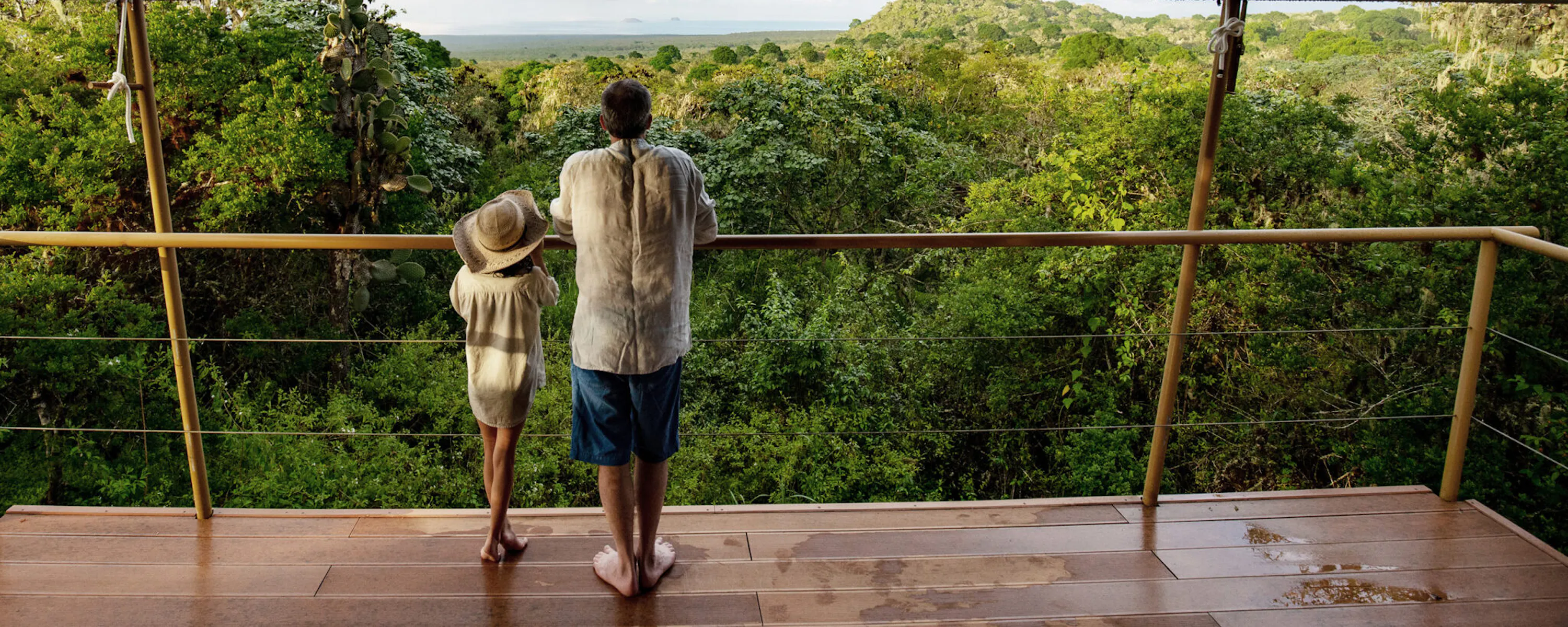 Two people stand at the opening of a safari-style tent, looking out over green hills beneath a wide cloudy sky.
