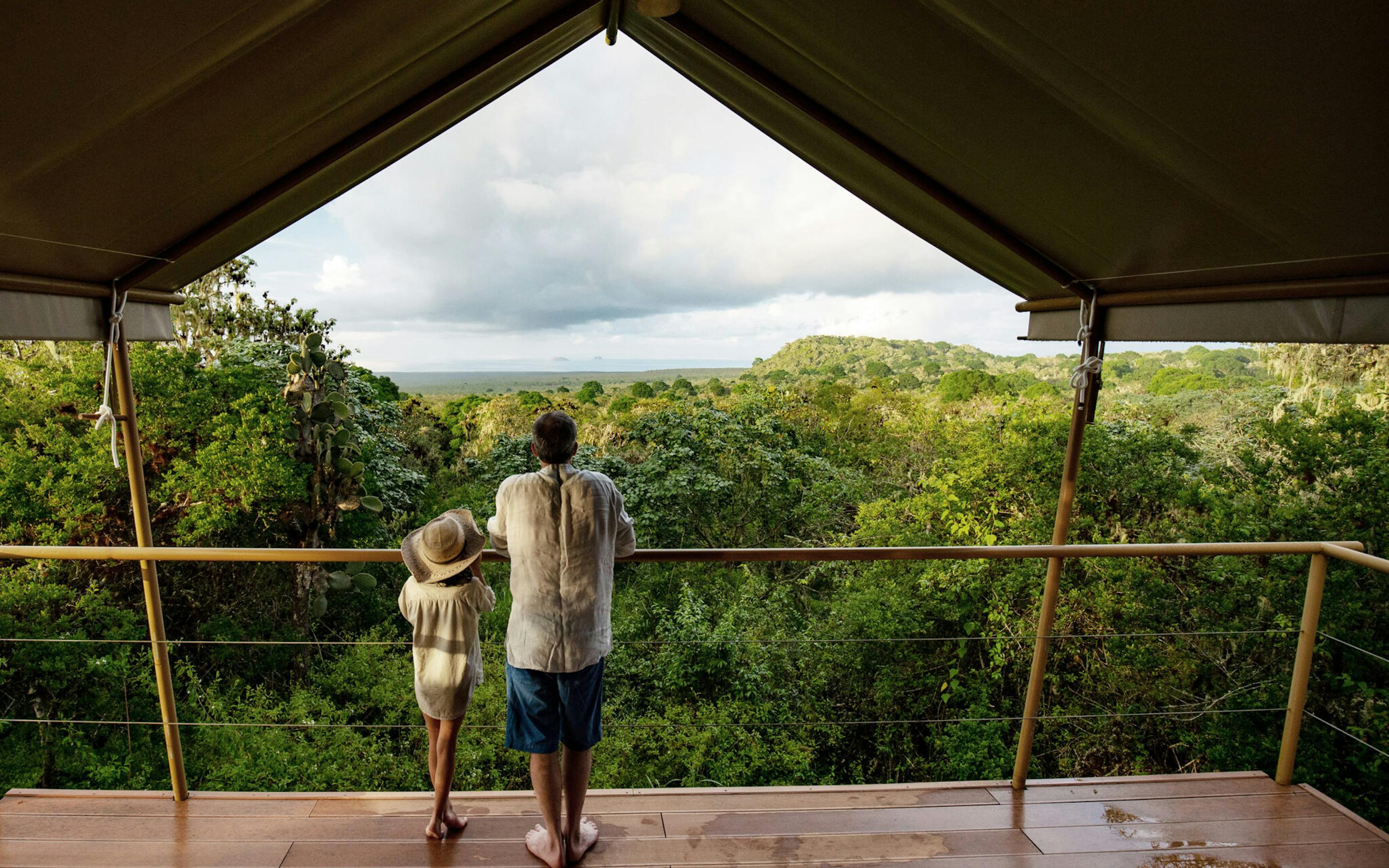 Two people stand at the opening of a safari-style tent, looking out over green hills beneath a wide cloudy sky.