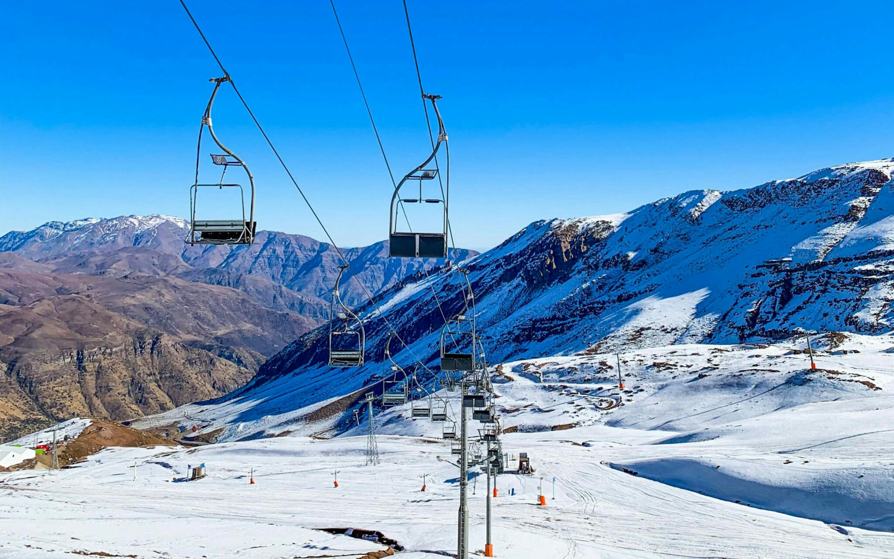 Empty ski chairs glide above snowy runs at Valle Nevado, framed by distant peaks and a deep, clear blue sky.