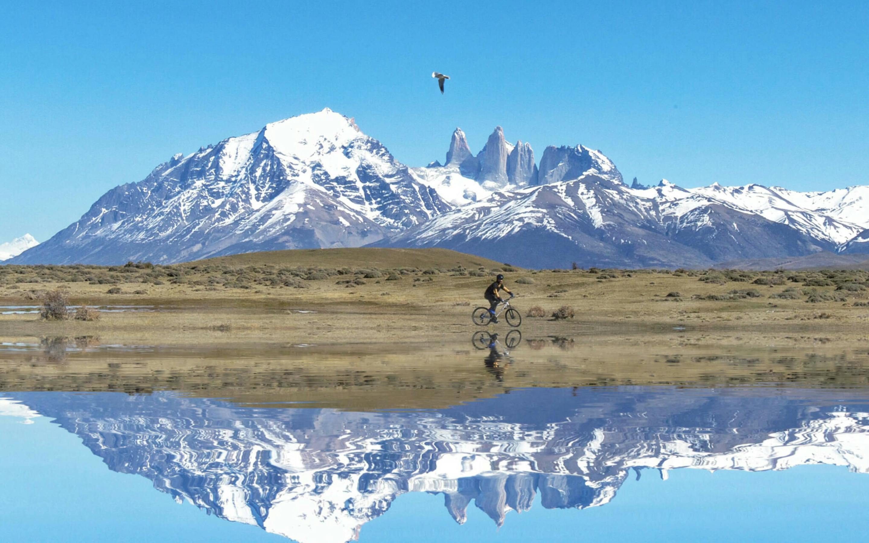 Snow-dusted Torres del Paine peaks reflect in a still lake, while small birds dot the foreground quiet shore and cyclist enjoys the views