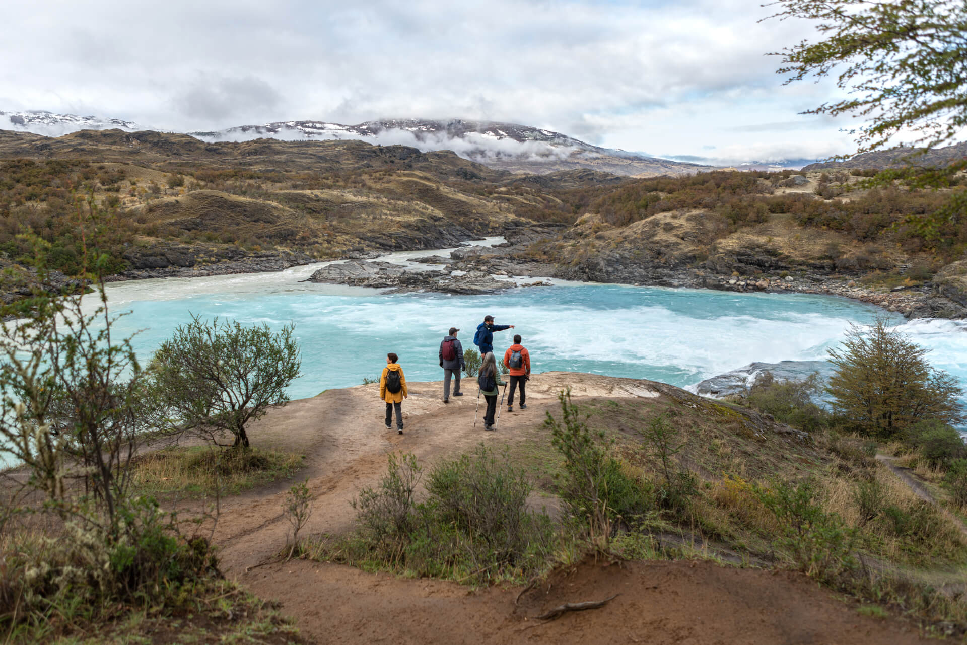 Hikers follow a lakeside trail through scrubby hills, with a pale blue lagoon and rugged peaks under cloud.