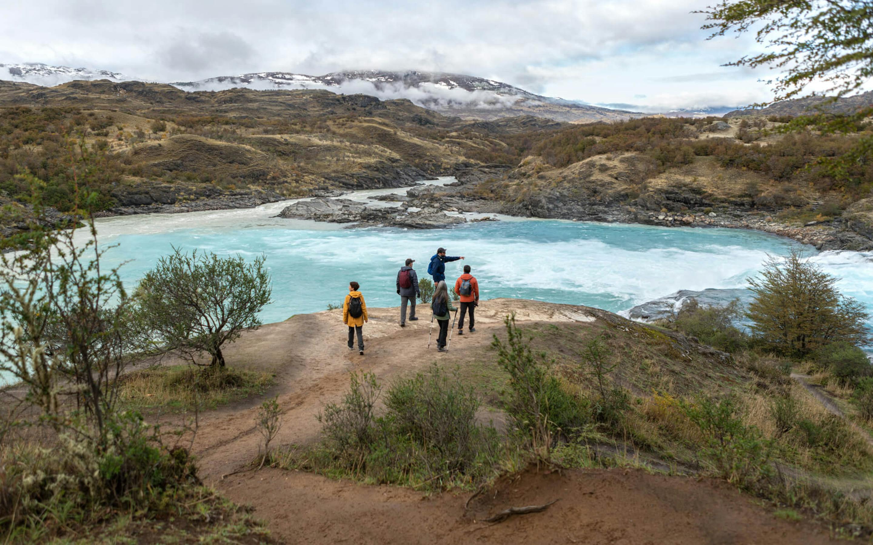 Hikers follow a lakeside trail through scrubby hills, with a pale blue lagoon and rugged peaks under cloud.