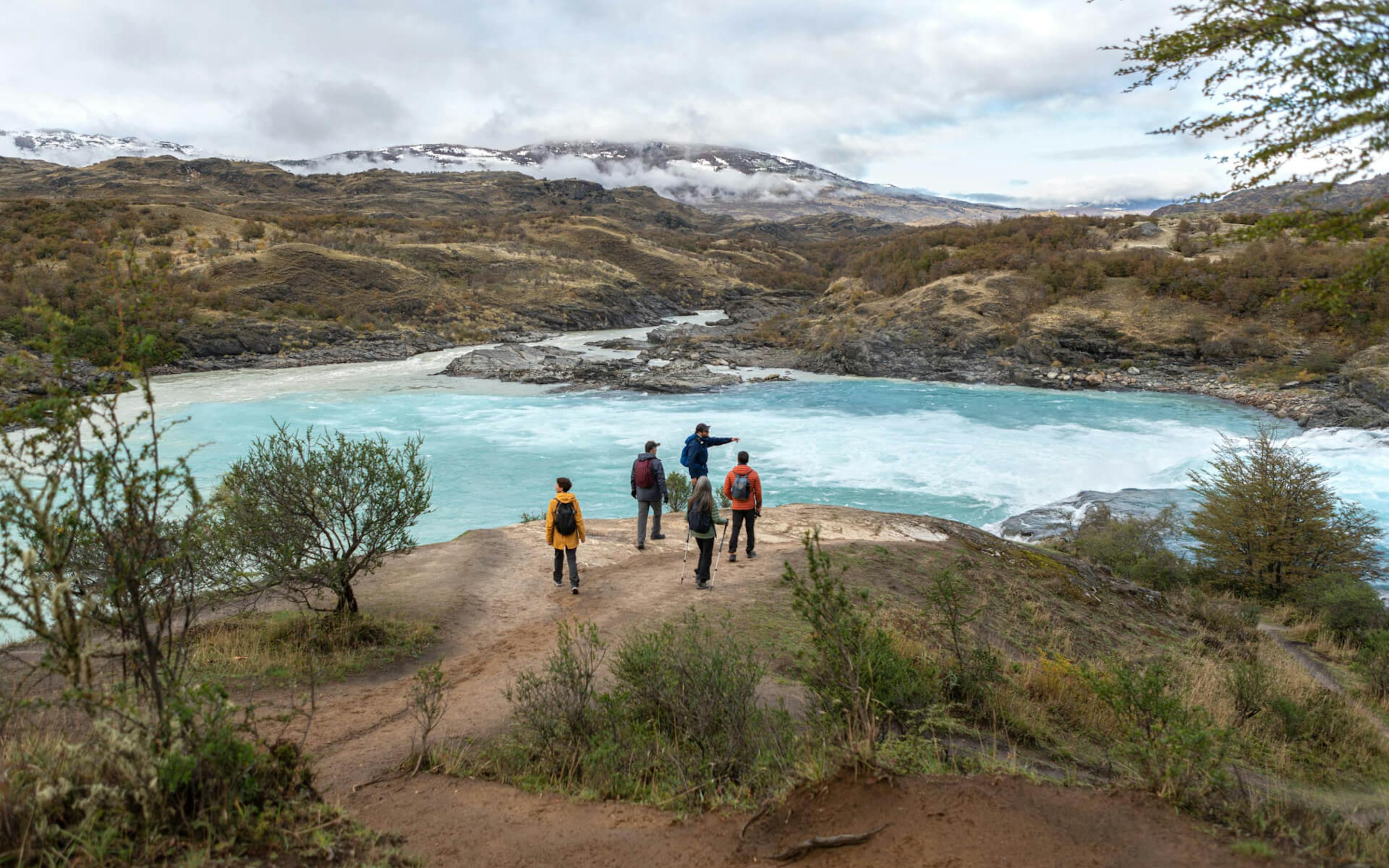 Hikers follow a lakeside trail through scrubby hills, with a pale blue lagoon and rugged peaks under cloud.