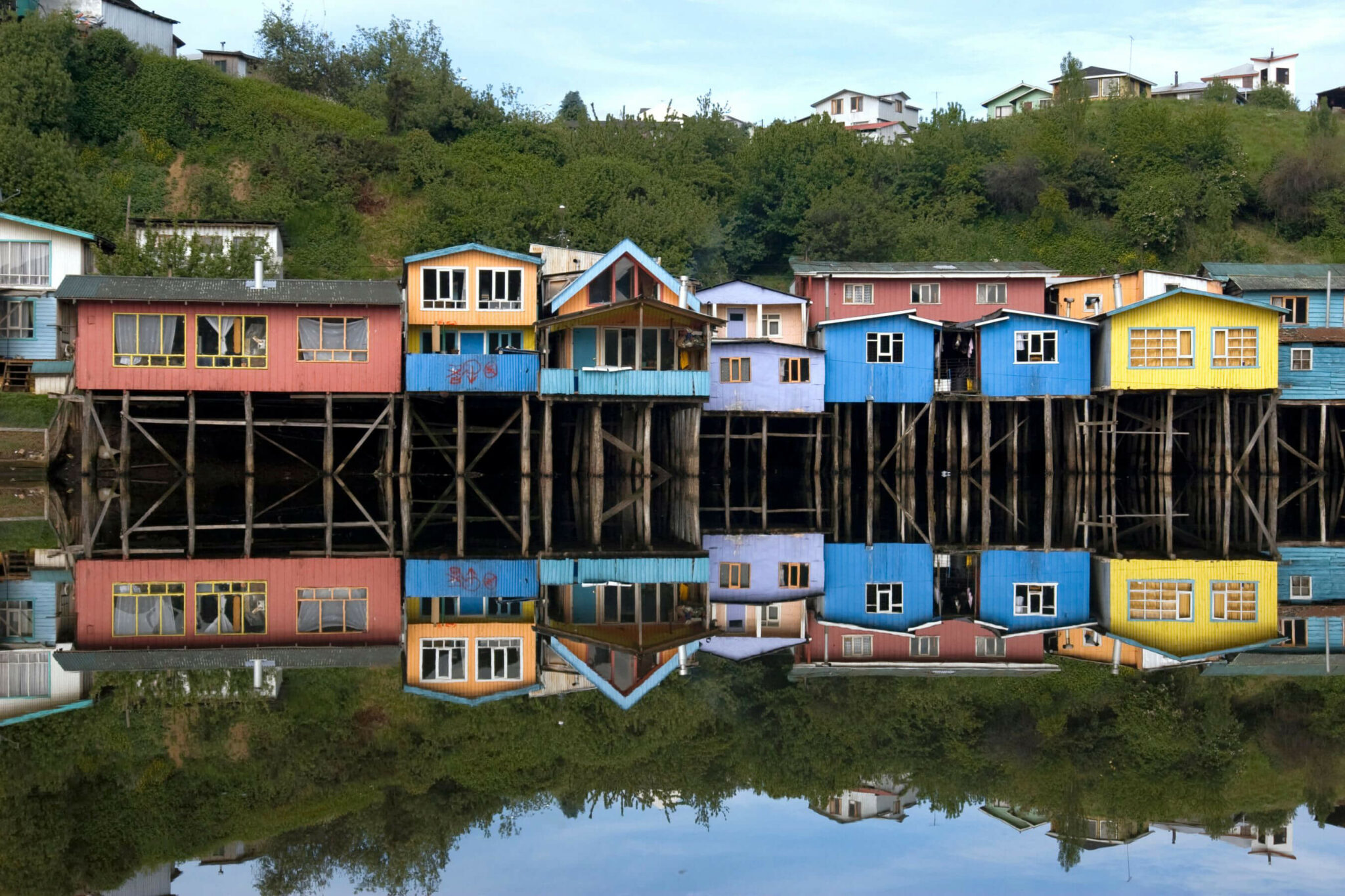 Bright stilt houses line the waterfront in Chiloé, mirrored in calm water beneath a soft, pale evening sky.