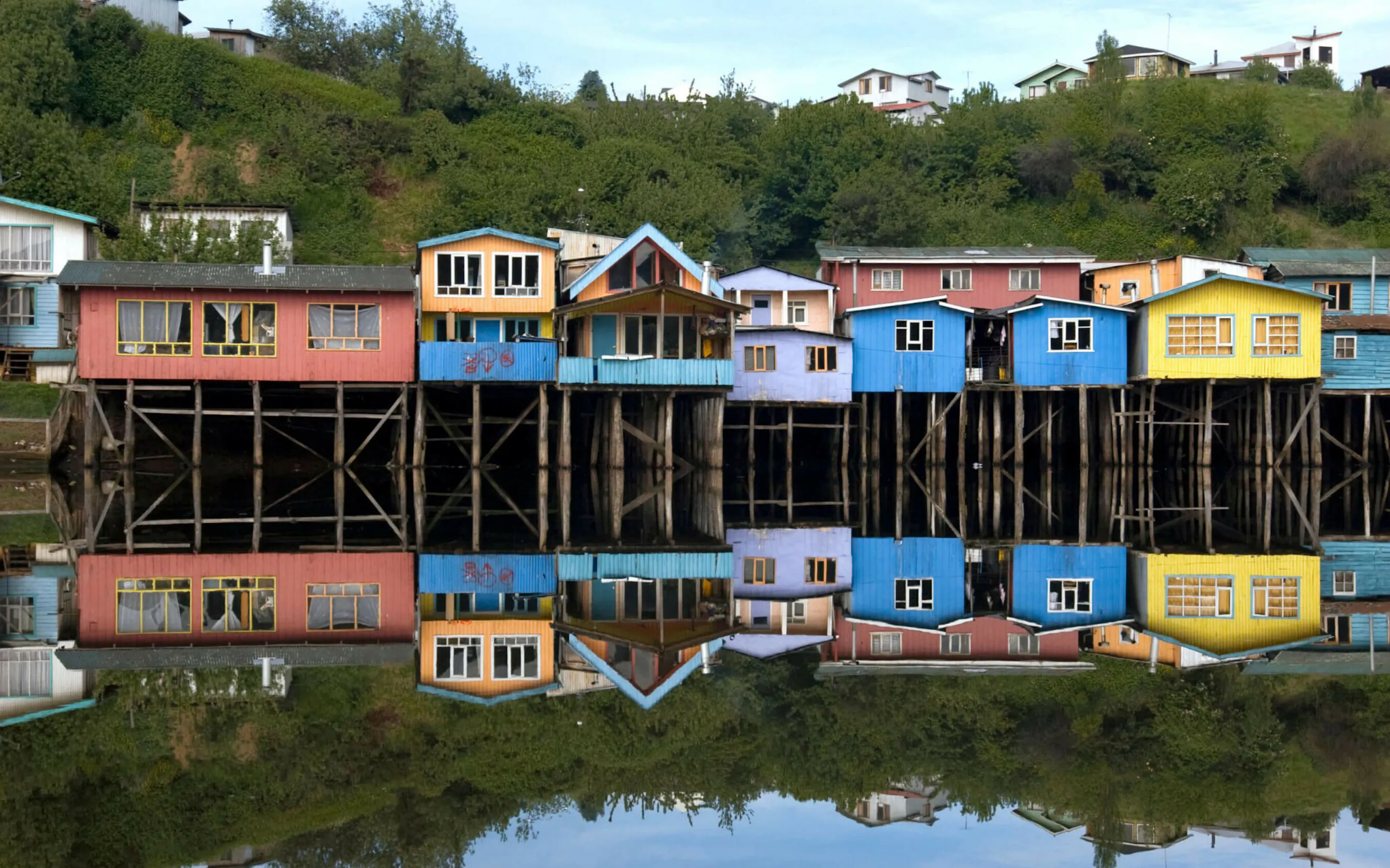 Bright stilt houses line the waterfront in Chiloé, mirrored in calm water beneath a soft, pale evening sky.