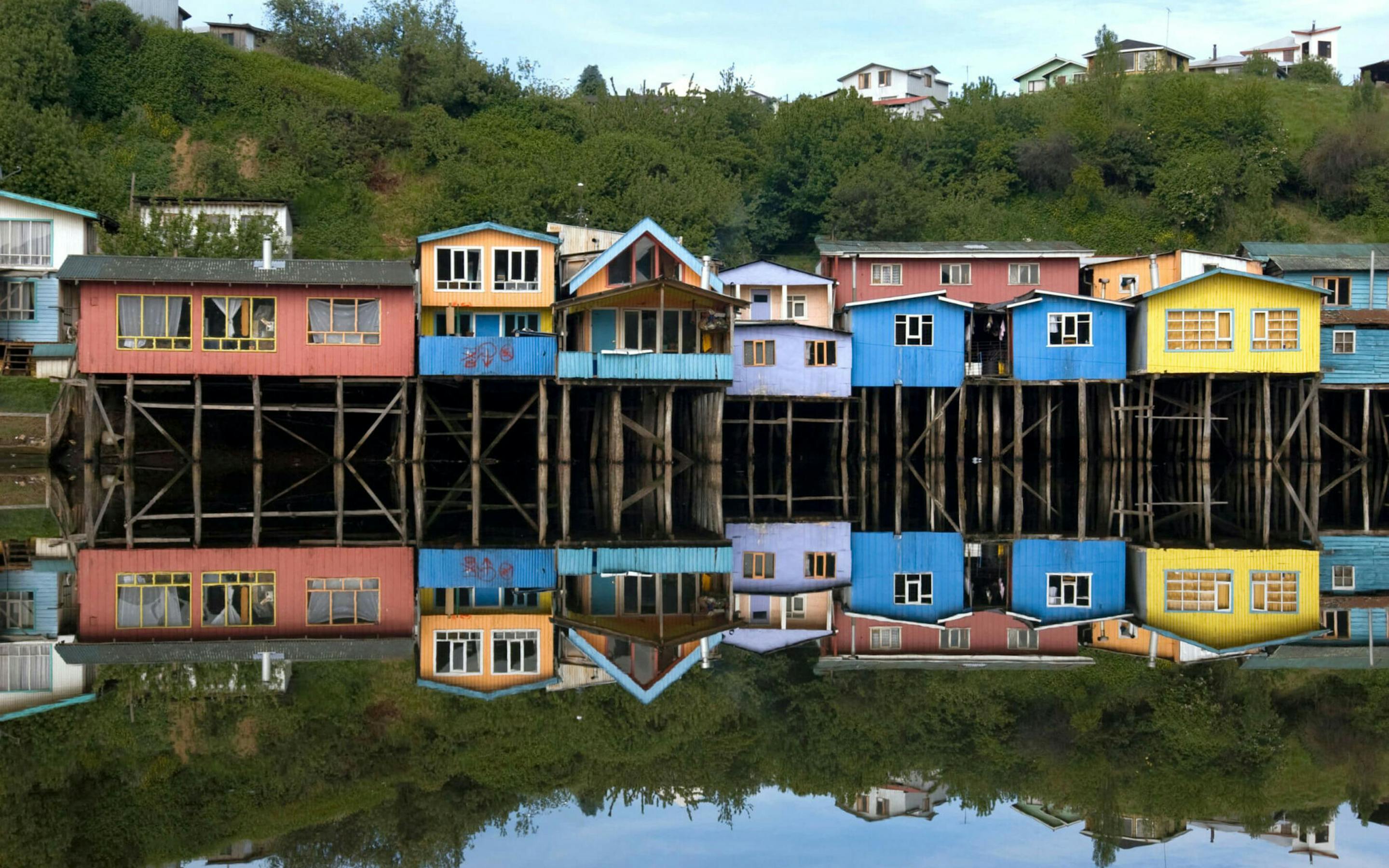 Bright stilt houses line the waterfront in Chiloé, mirrored in calm water beneath a soft, pale evening sky.