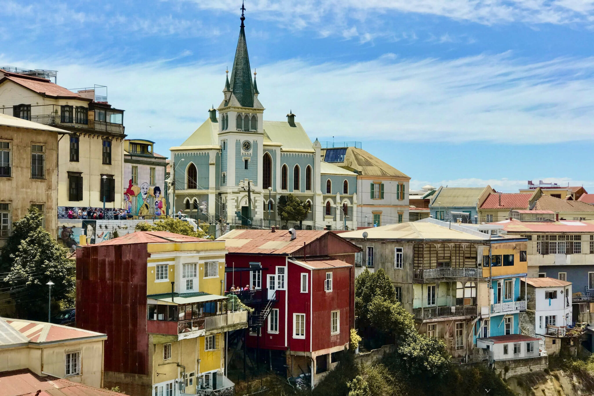Colorful hillside homes and a church steeple rise above the waterfront, with stacked rooftops in bright sun.