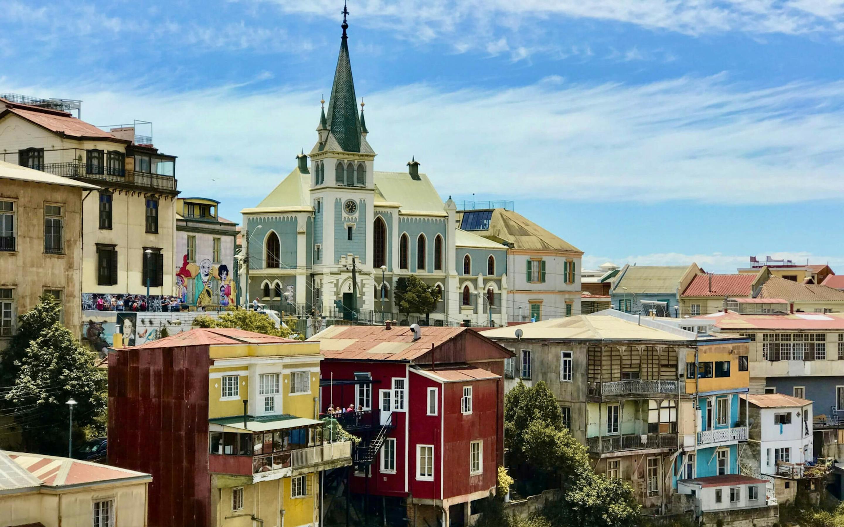 Colorful hillside homes and a church steeple rise above the waterfront, with stacked rooftops in bright sun.