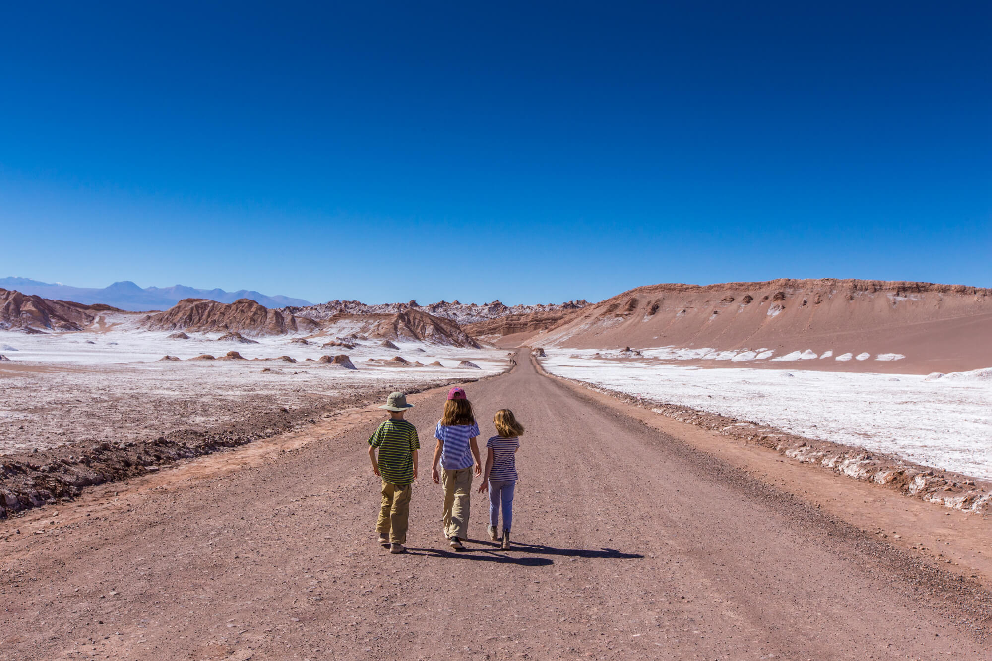 Three travelers walk down a wide dirt road across the Atacama desert, with salt flats and red hills ahead.