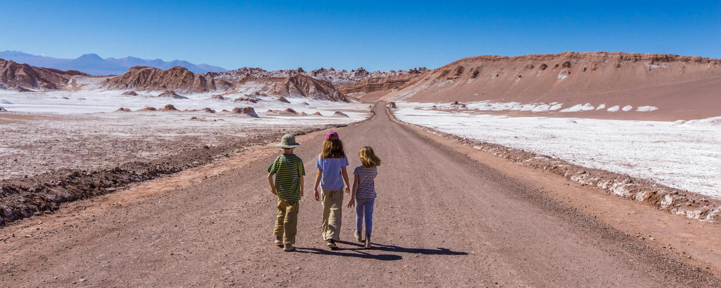 Three travelers walk down a wide dirt road across the Atacama desert, with salt flats and red hills ahead.