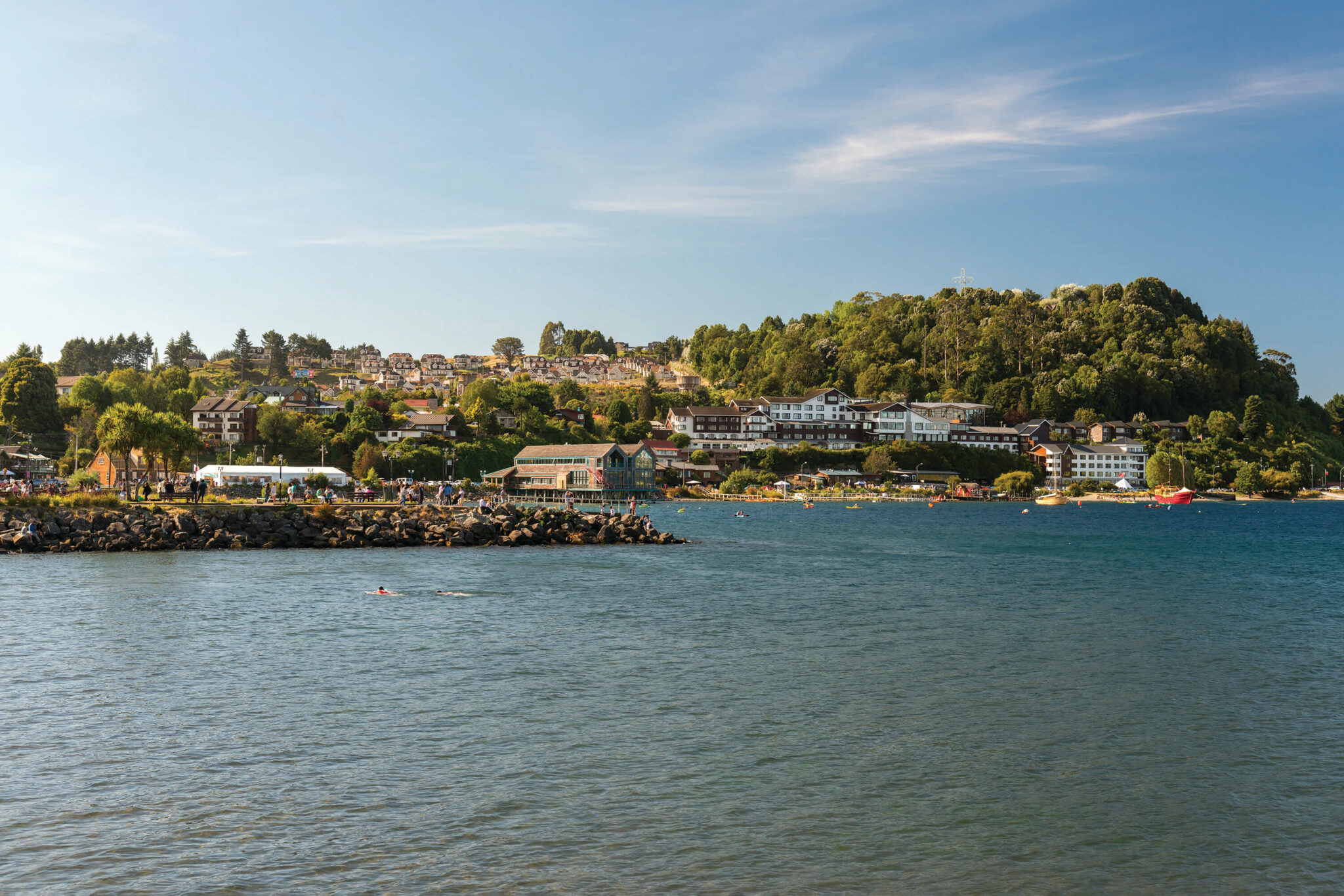 Houses and small boats line a sheltered bay, with forested hills rising behind the waterfront under a clear sky.