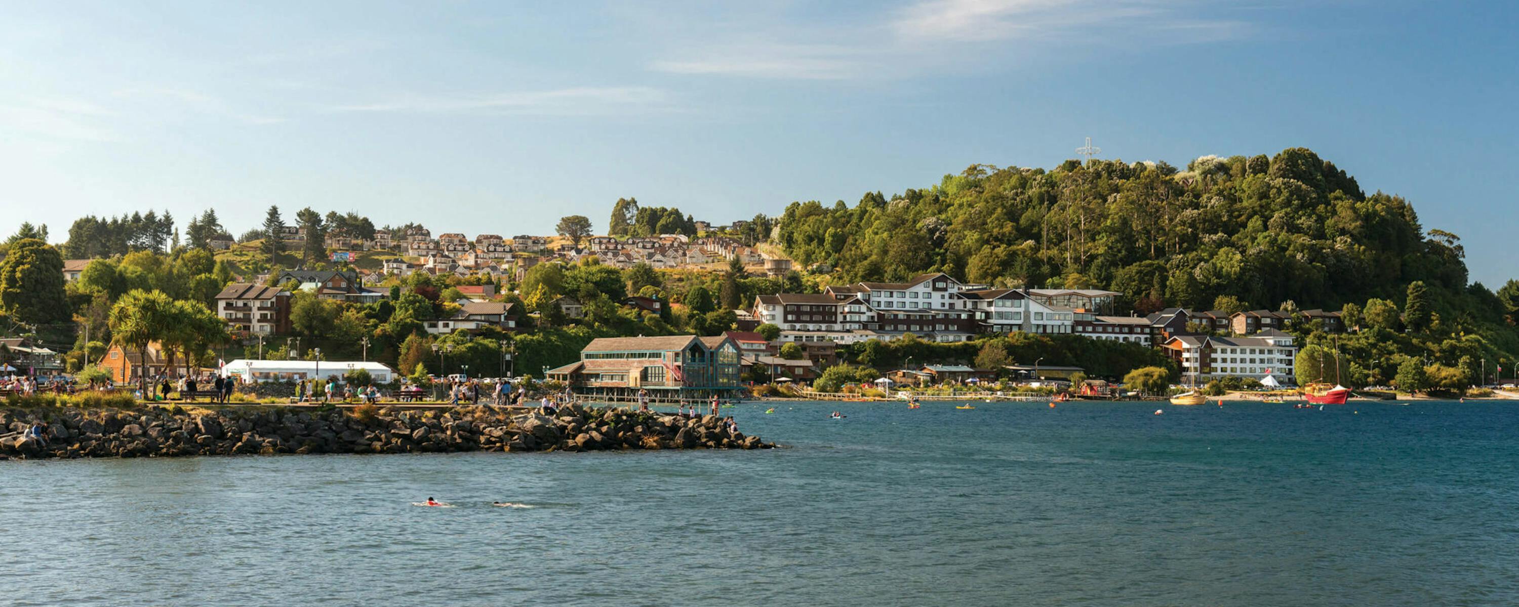 Houses and small boats line a sheltered bay, with forested hills rising behind the waterfront under a clear sky.