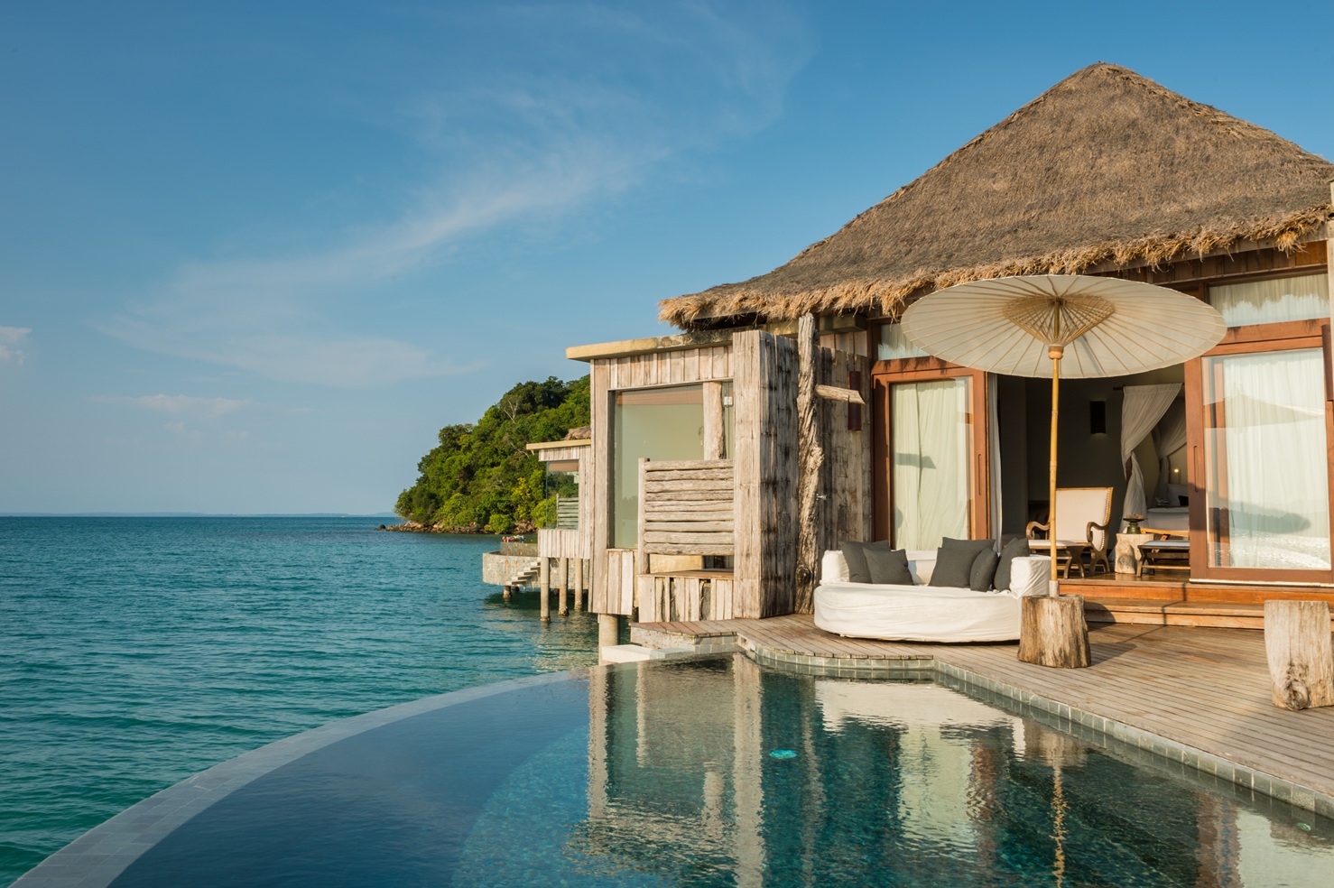 An overwater villa opens to an infinity pool above the sea, with a thatched roof and lounge seating at the edge.