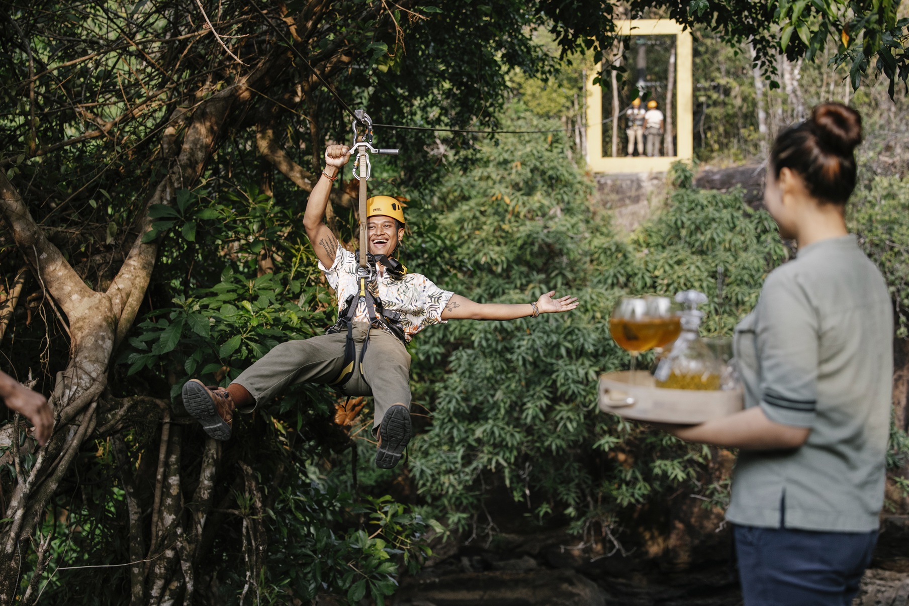 A helmeted guest on a zip line reaches for a drink offered on a tray, surrounded by dense jungle foliage.
