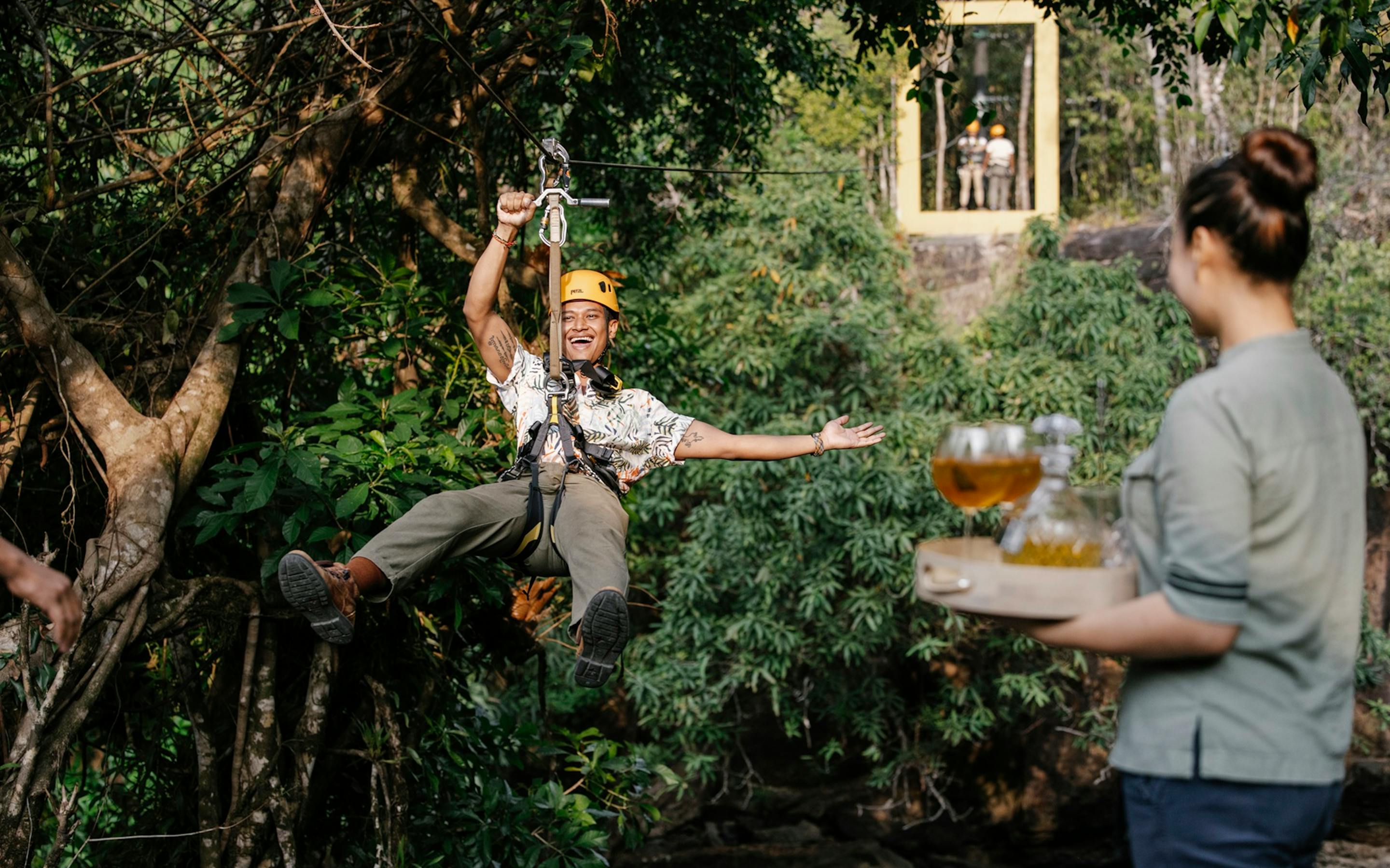 A helmeted guest on a zip line reaches for a drink offered on a tray, surrounded by dense jungle foliage.