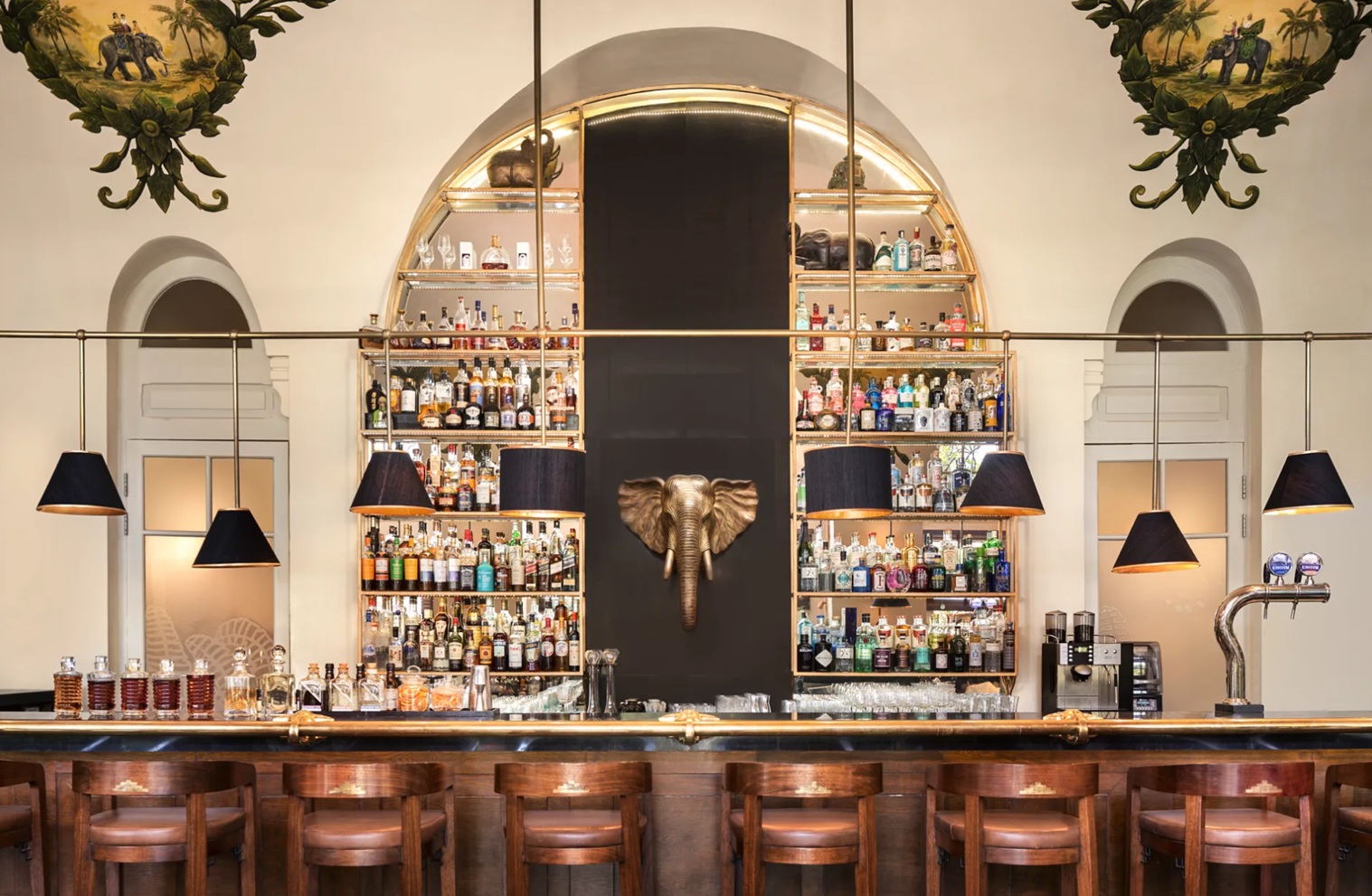 A classic bar interior features arched mirrors, glass shelves of bottles, and wooden stools under warm pendant lights.