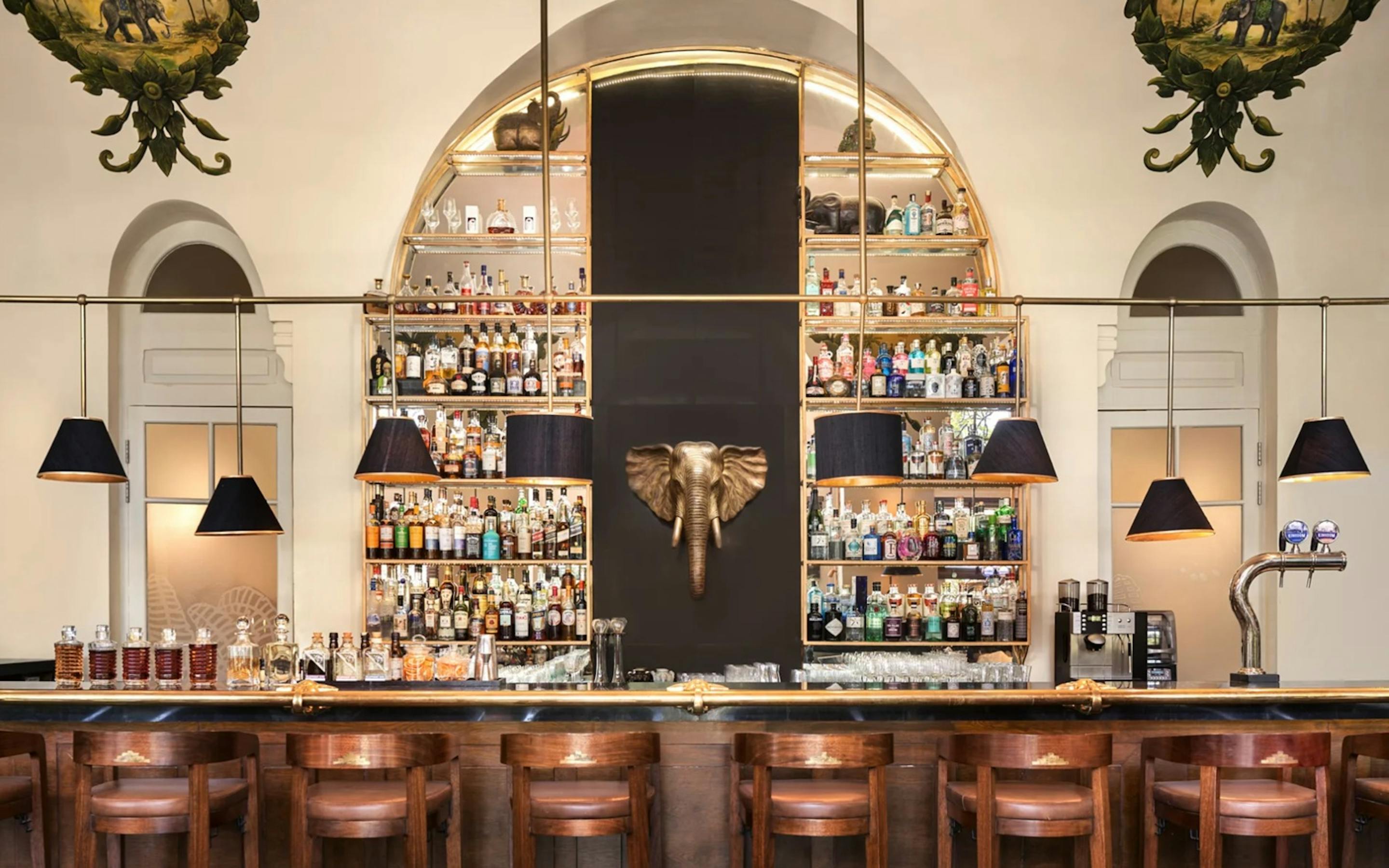 A classic bar interior features arched mirrors, glass shelves of bottles, and wooden stools under warm pendant lights.