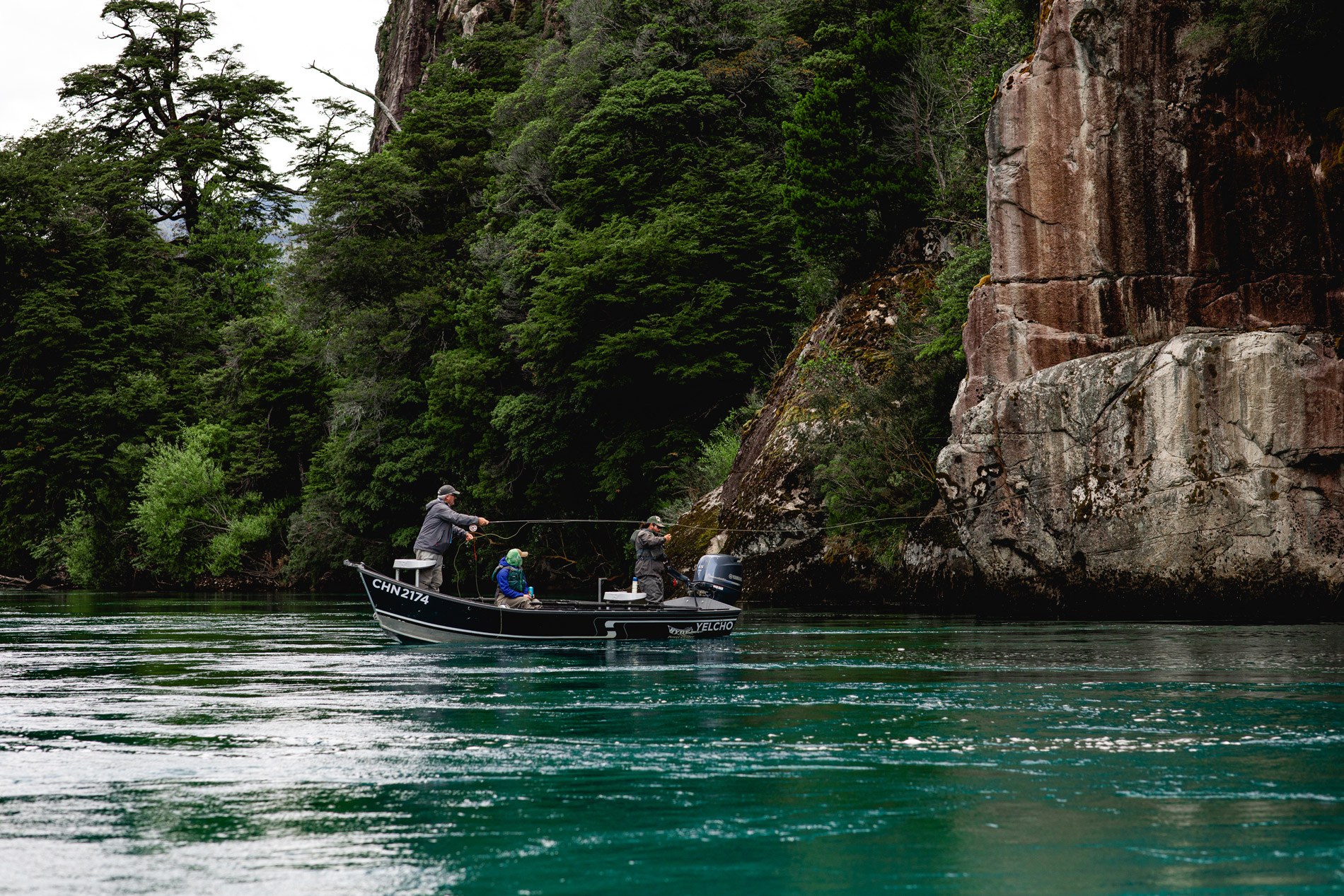 Small boat glides across a turquoise lake beside tall rock cliffs and dense green forest along the shoreline.