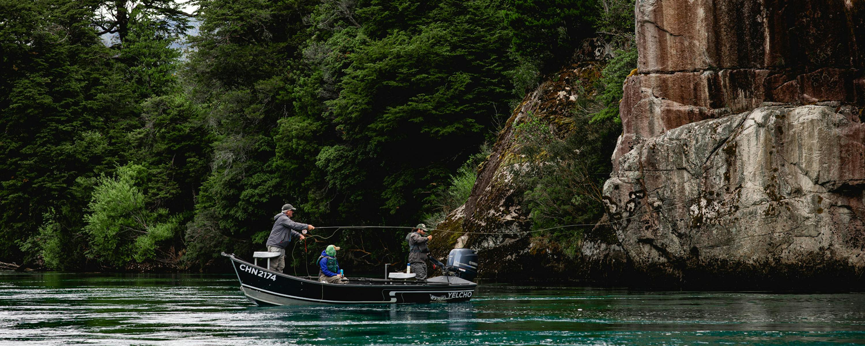 Small boat glides across a turquoise lake beside tall rock cliffs and dense green forest along the shoreline.