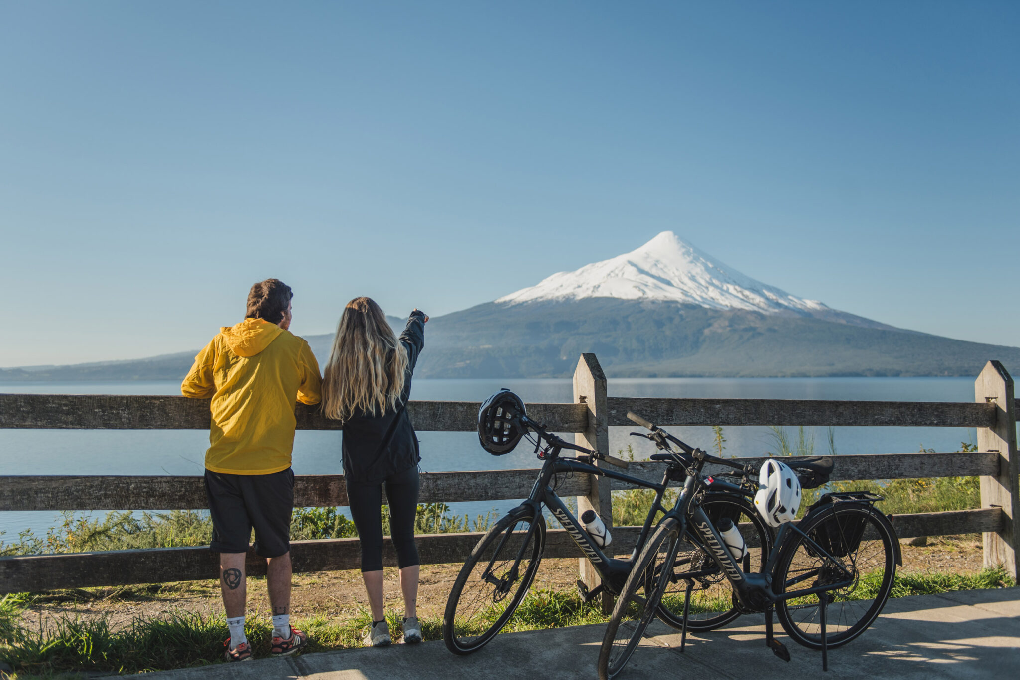 Two travelers stand by bicycles on a lakeside viewpoint, looking toward a conical volcano across calm water.
