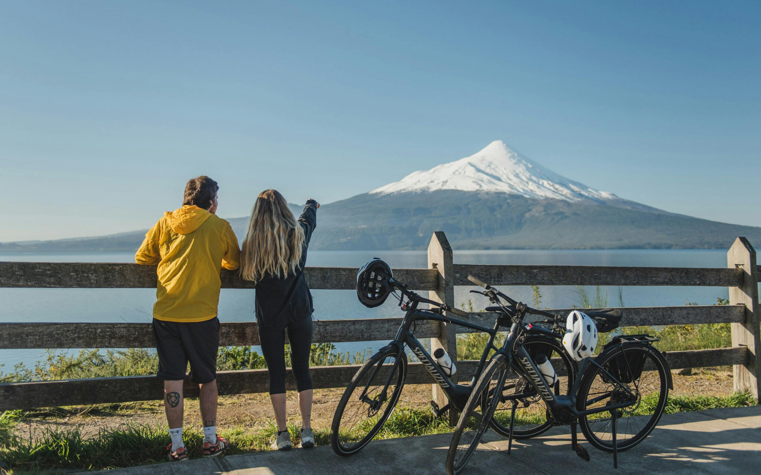 Two travelers stand by bicycles on a lakeside viewpoint, looking toward a conical volcano across calm water.