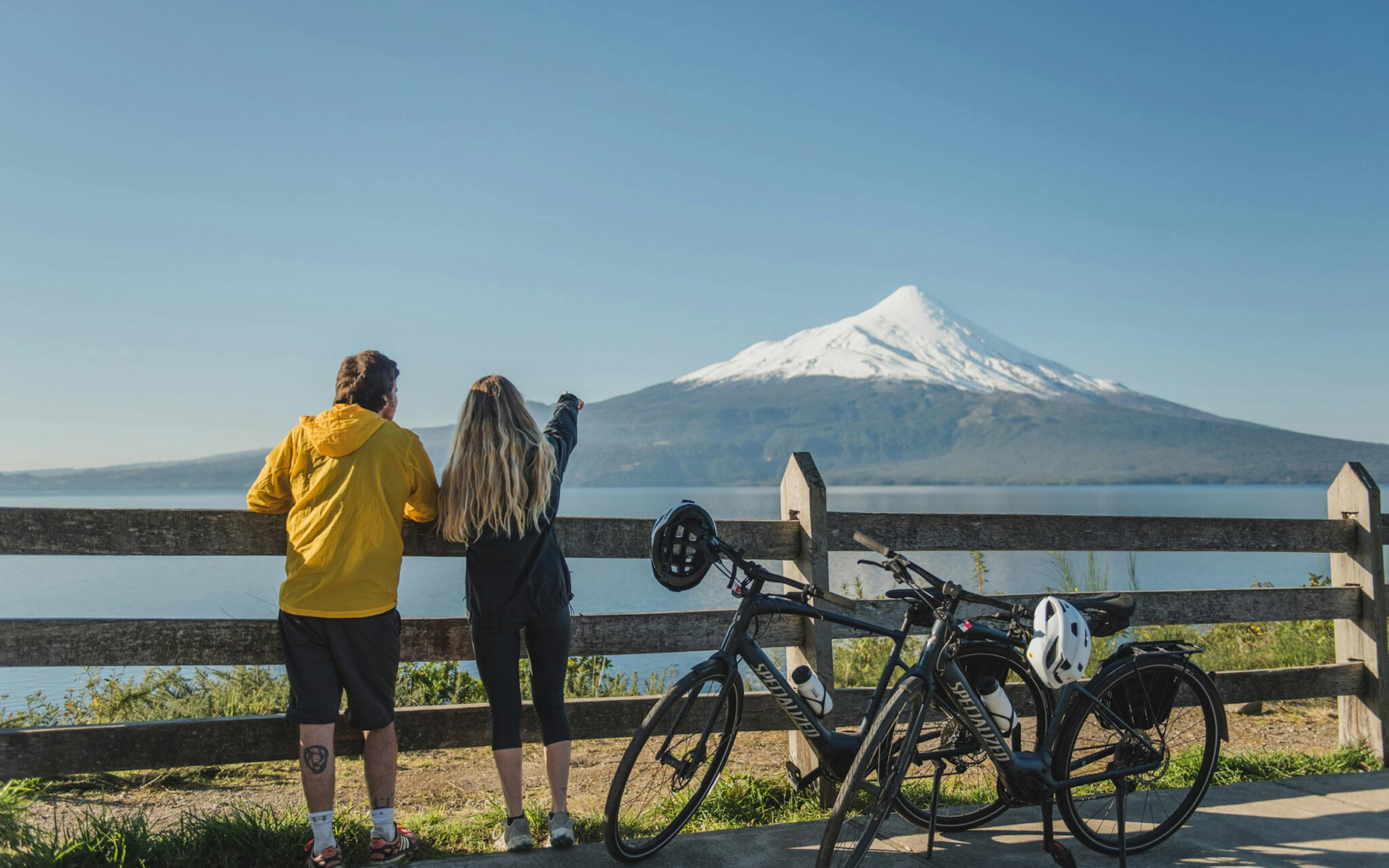 Two travelers stand by bicycles on a lakeside viewpoint, looking toward a conical volcano across calm water.